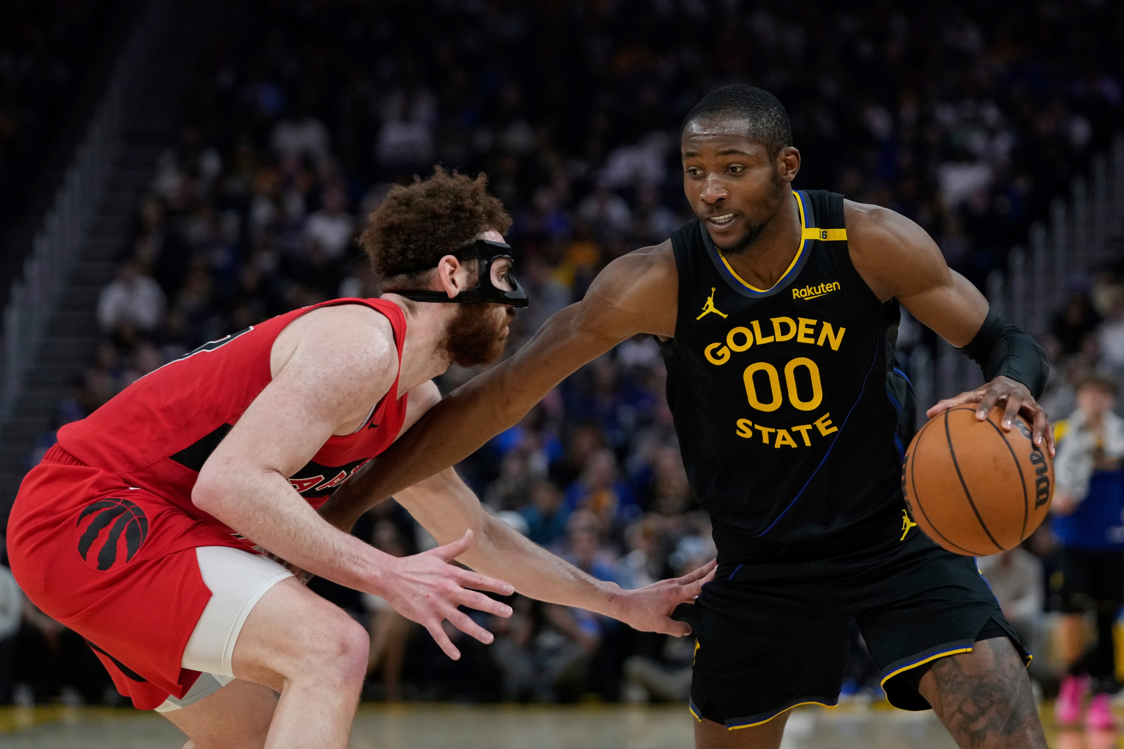 A basketball player in a Golden State uniform dribbles while being guarded by a player in a red uniform with a black face mask.