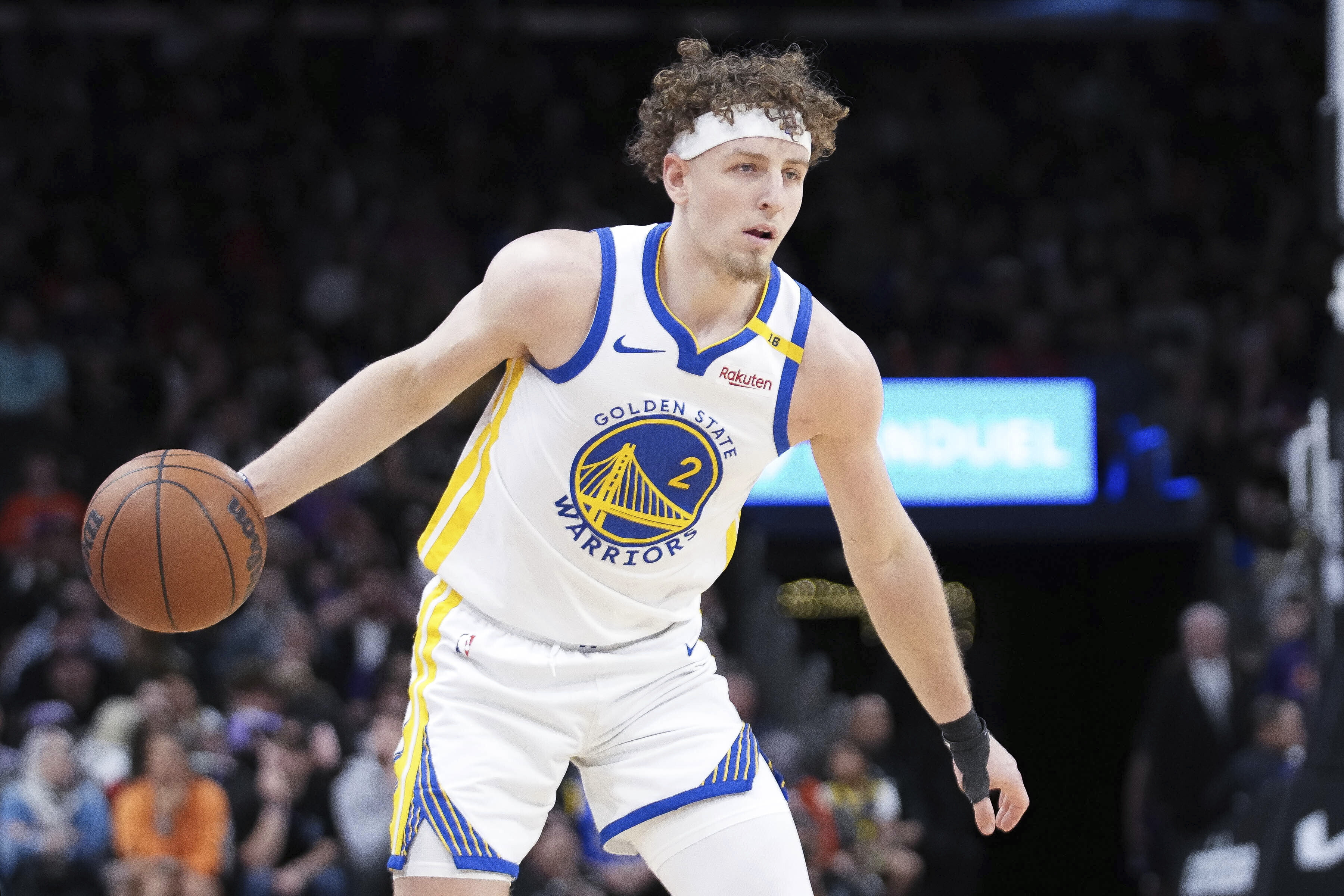 A basketball player in a Golden State Warriors uniform dribbles the ball on the court. He's wearing a white jersey with blue and yellow trim and a headband.