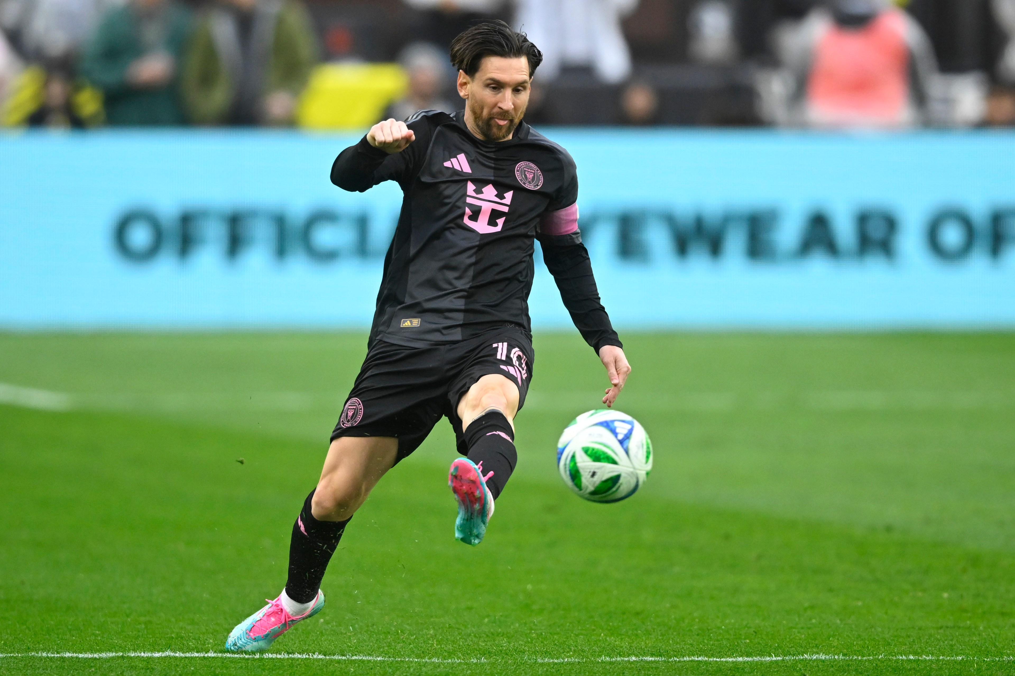 A soccer player in a black and pink uniform kicks a ball on a grassy field. The background is a blurred advertisement and spectators.