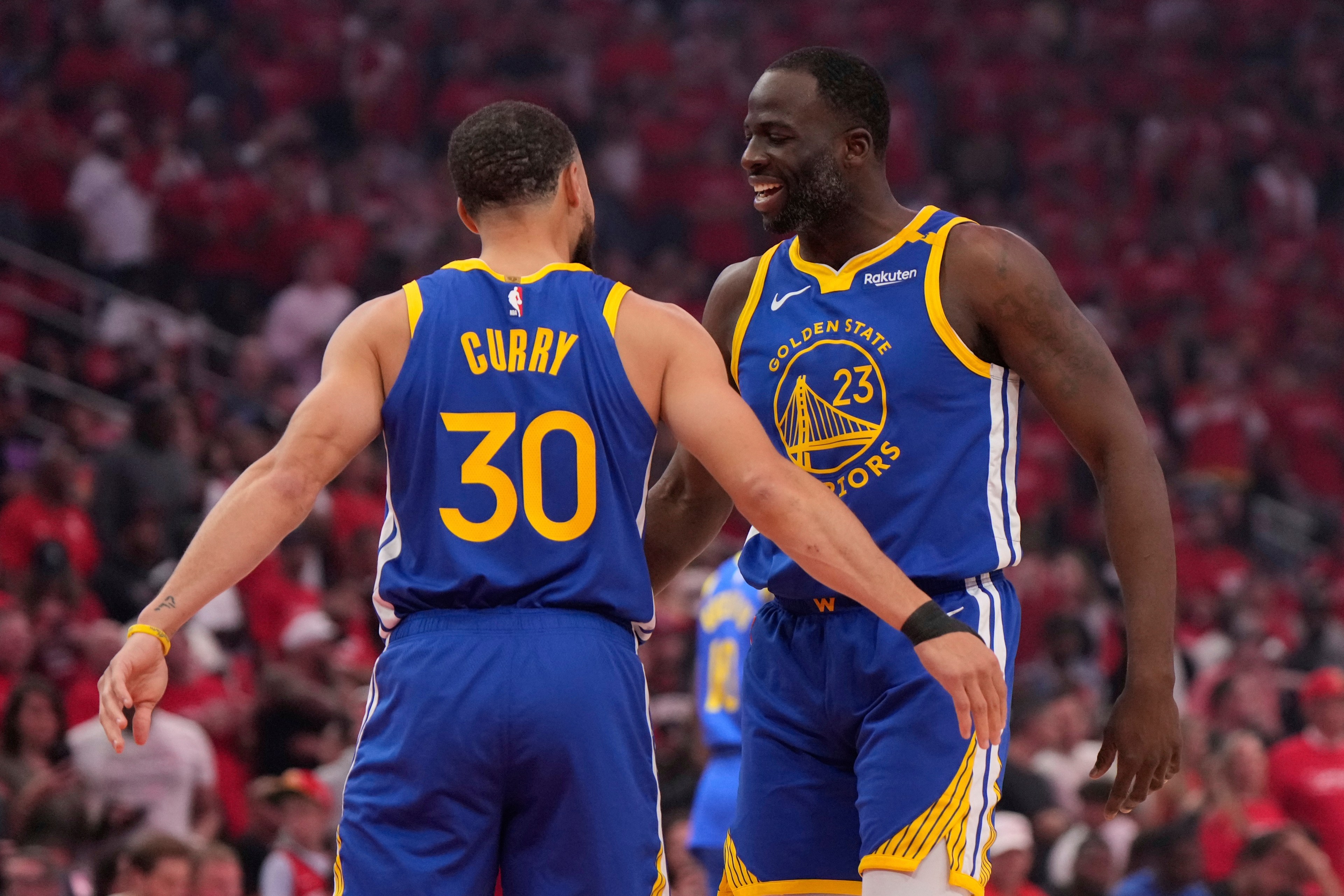 Two basketball players in blue Golden State Warriors jerseys are on a court, embracing and smiling. One jersey shows the name "Curry" and number 30.