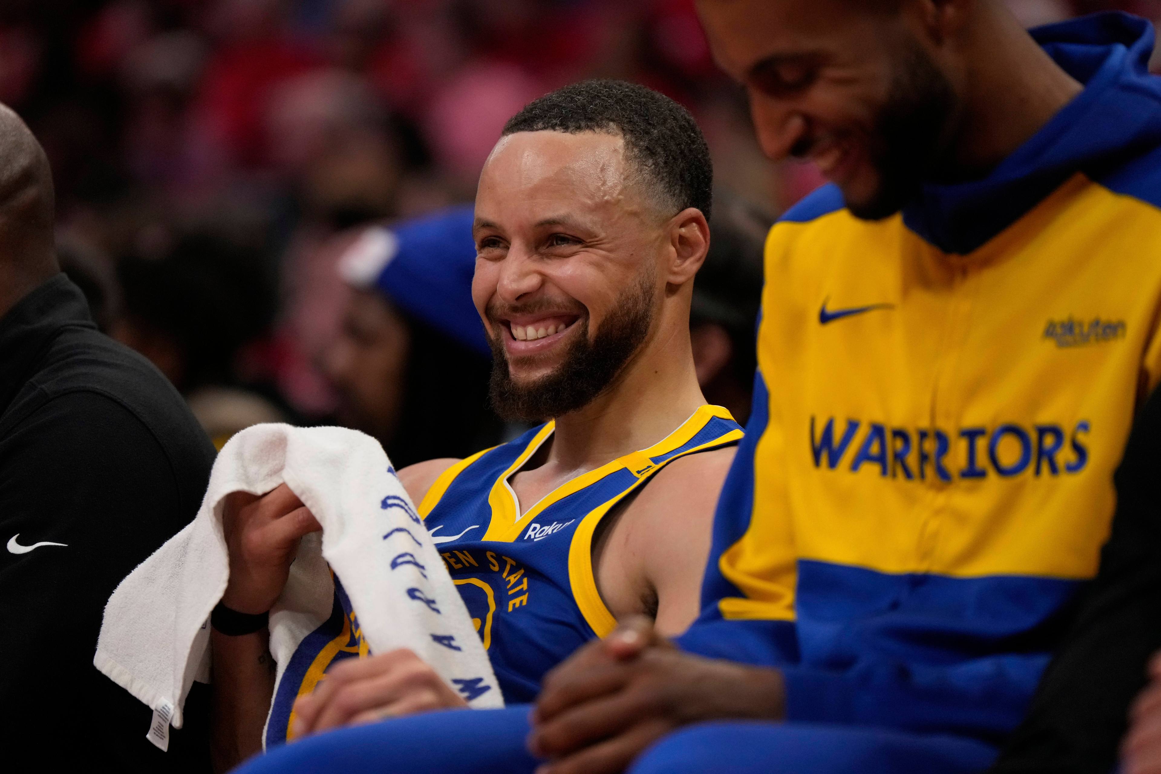 A basketball player in a Warriors jersey smiles broadly, holding a towel, while sitting on the bench. Another person next to him wears a matching yellow and blue jacket.