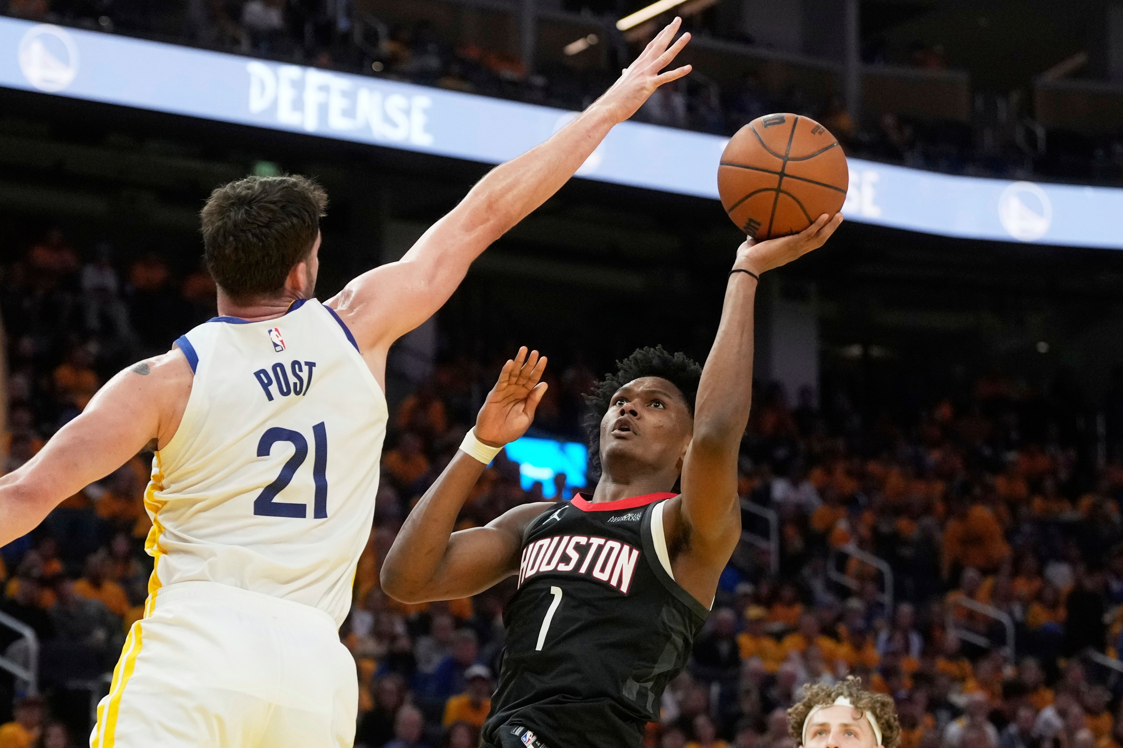 A basketball player in a black Houston jersey attempts a layup while a player in a white jersey labeled &quot;POST 21&quot; tries to block him. The crowd watches intensely.