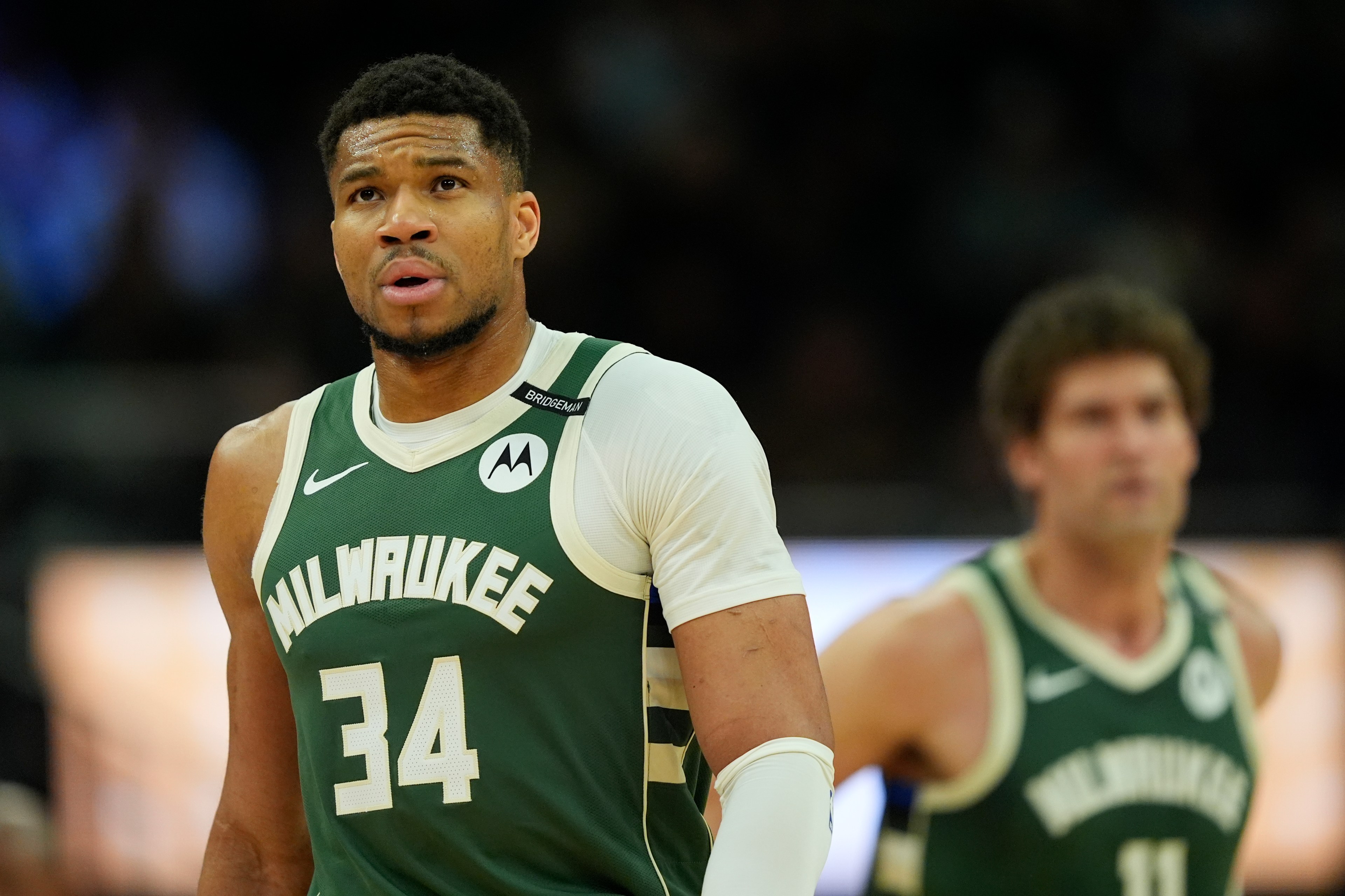 A basketball player in a green Milwaukee jersey with number 34 looks focused, while another player in the background is slightly out of focus.