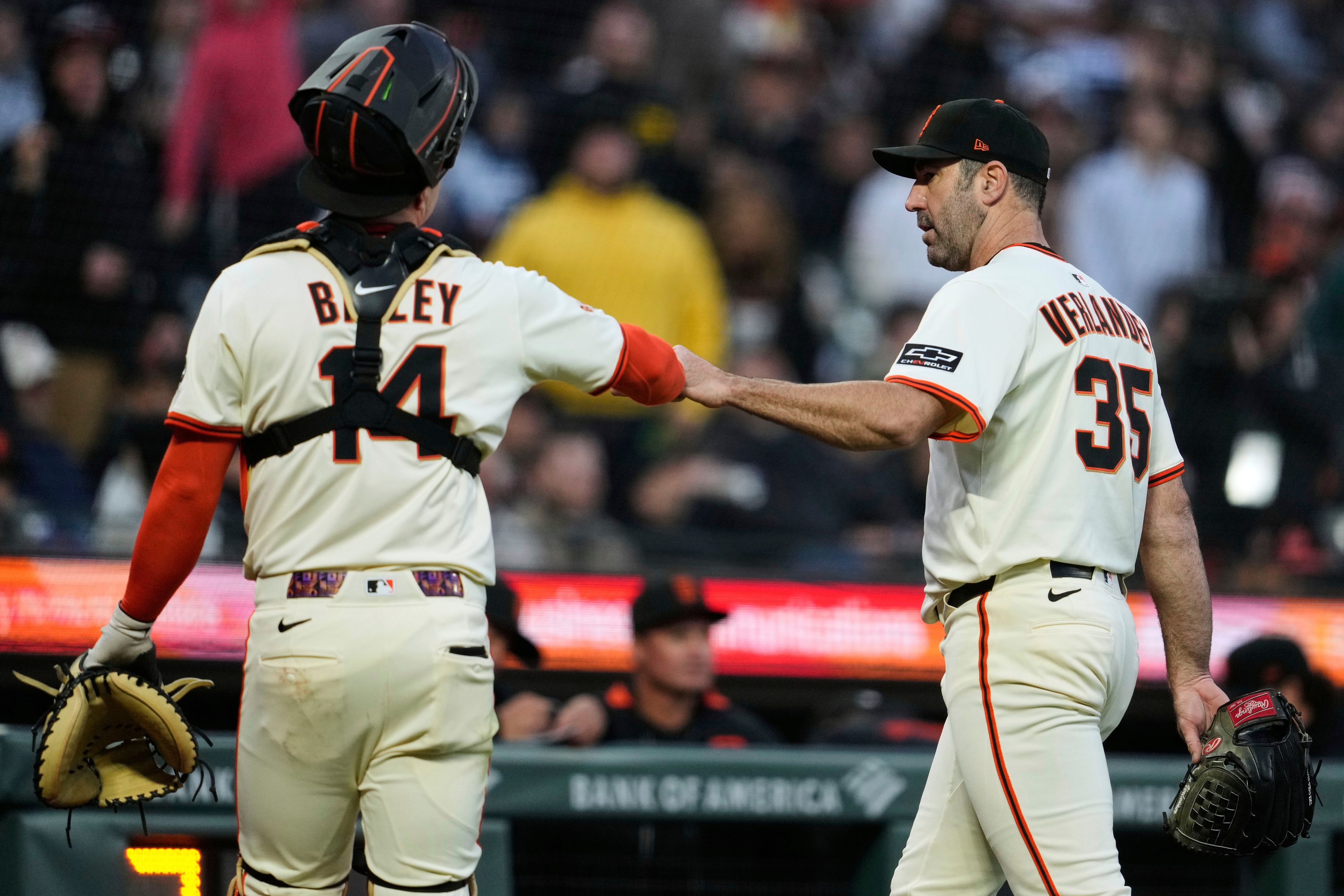 Two baseball players dressed in white uniforms are seen fist-bumping. One wears catcher's gear, and the other holds a glove. Their uniforms display the names Bailey and Velander.