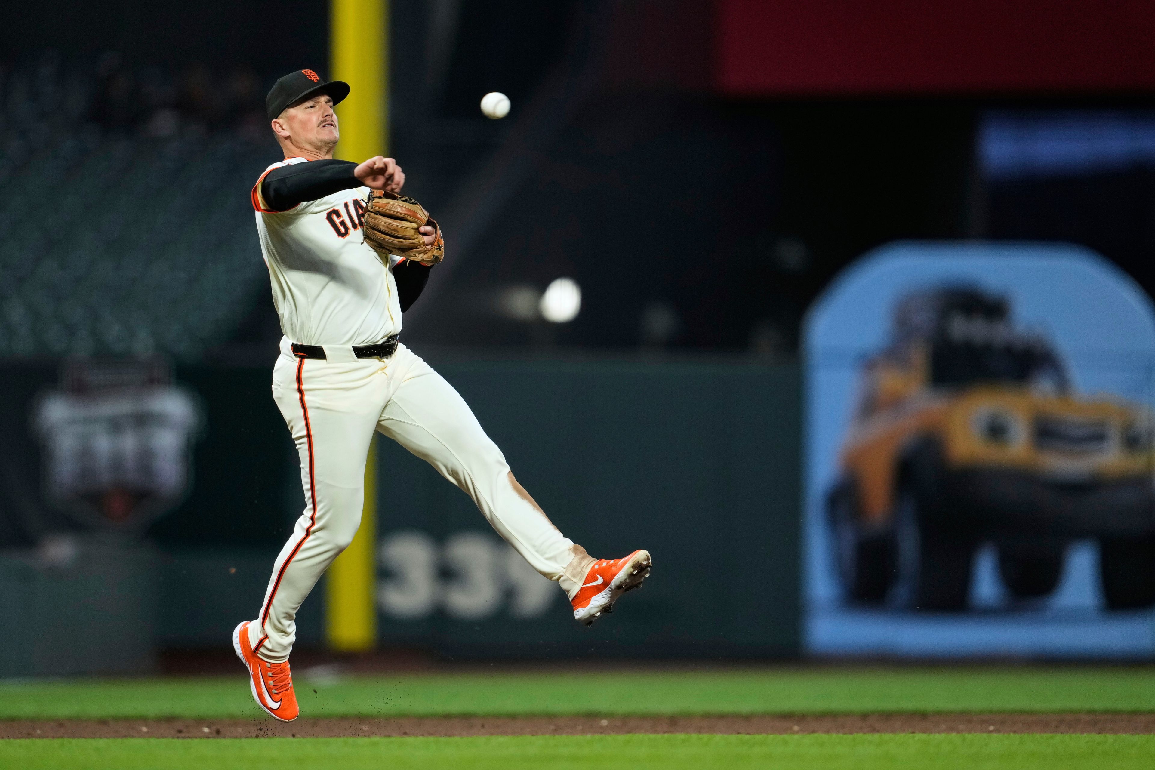 A baseball player in a Giants uniform is mid-action, airborne, throwing the ball with intensity. The background features stadium elements and a bright ad.