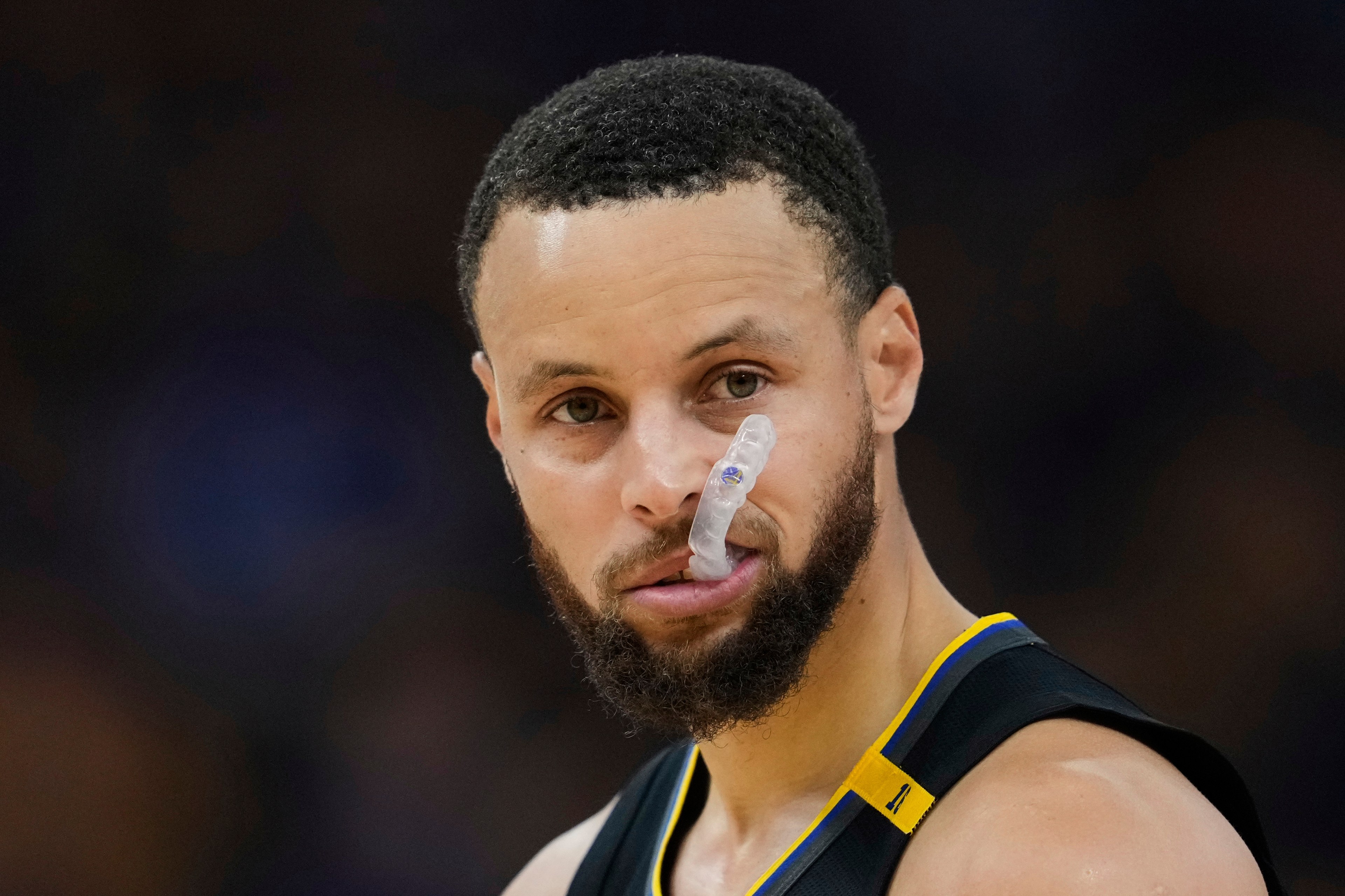 A basketball player with a beard is wearing a black jersey with yellow trim. He has a mouthguard hanging from his mouth, looking directly at the camera.