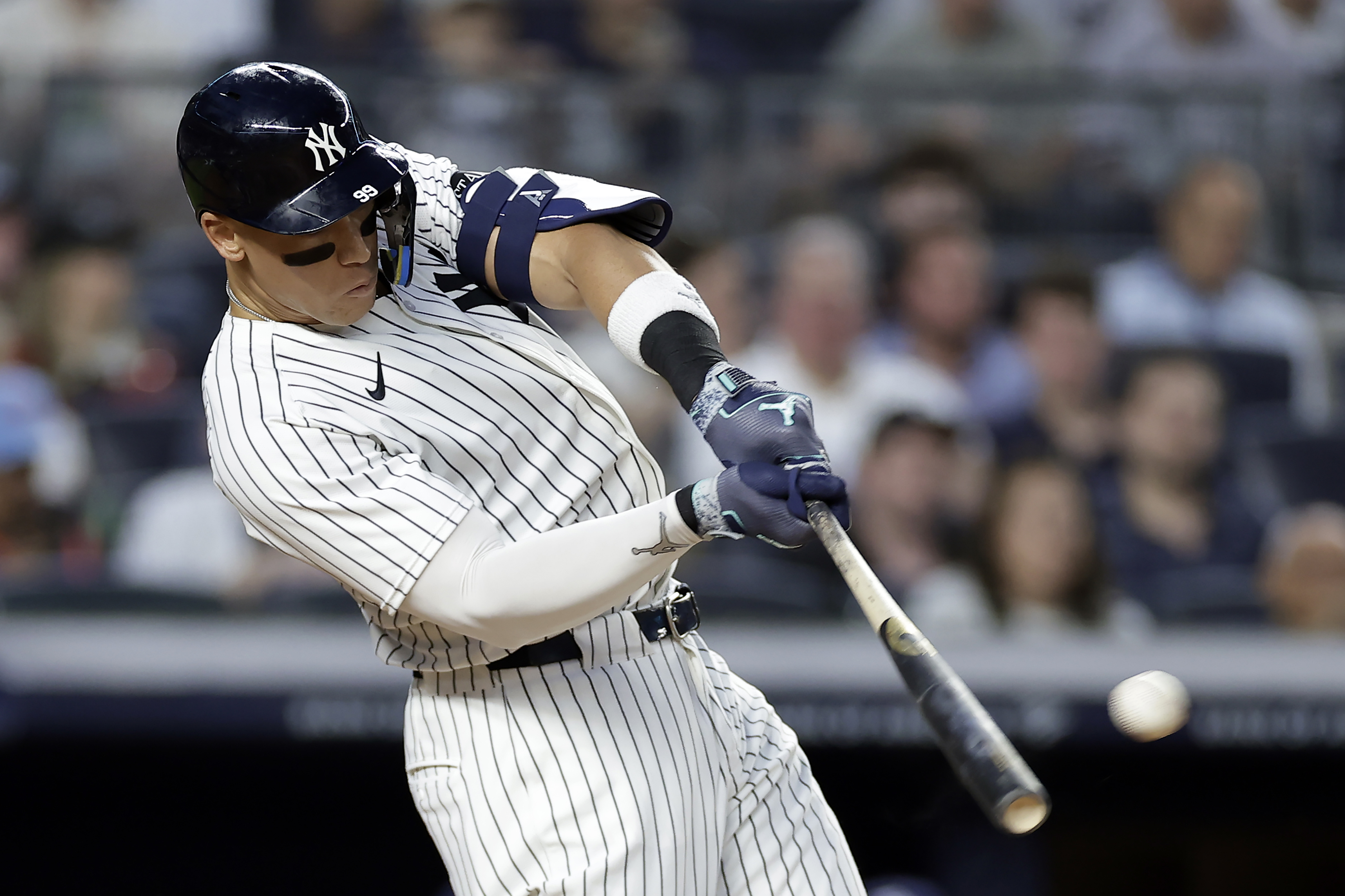 A baseball player in a pinstripe uniform powerfully swings a bat, hitting a baseball. He's focused, wearing a helmet and arm guard, with a blurred crowd in the background.