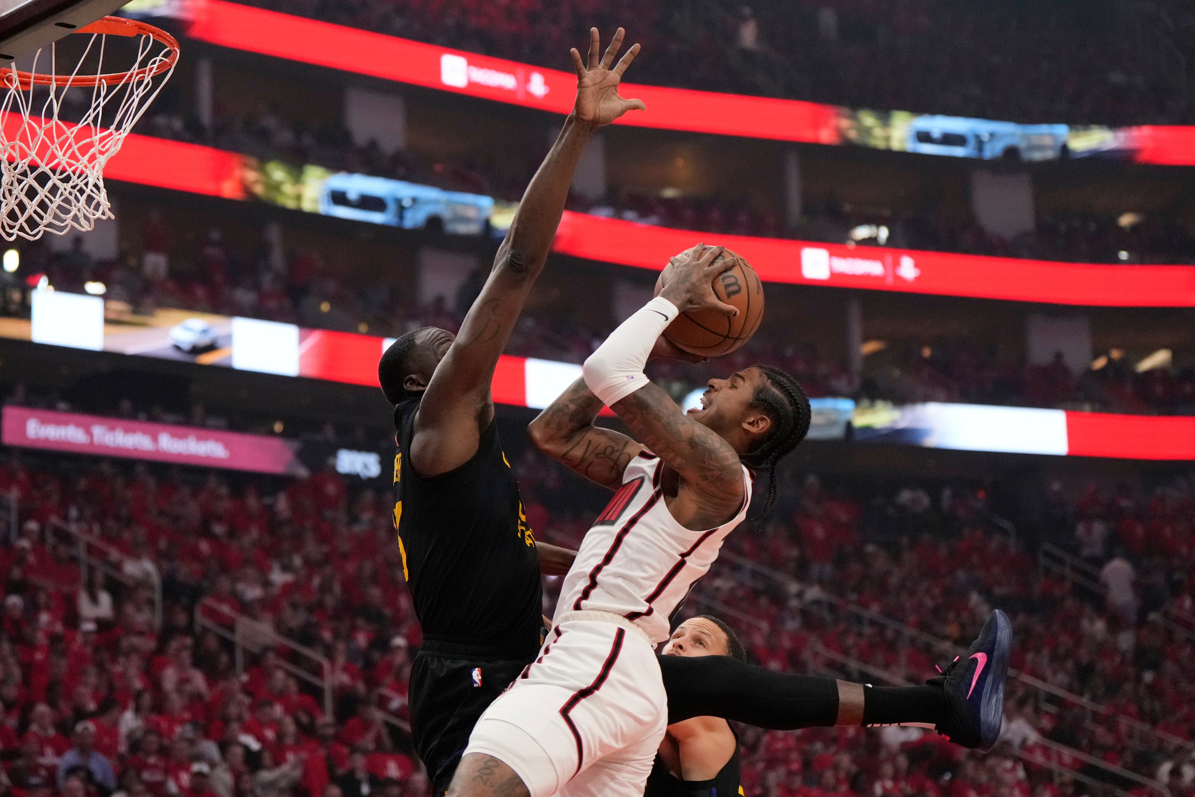 A basketball player in a white jersey jumps to shoot the ball, while a defender in a black jersey attempts to block him. The background shows a crowded arena.