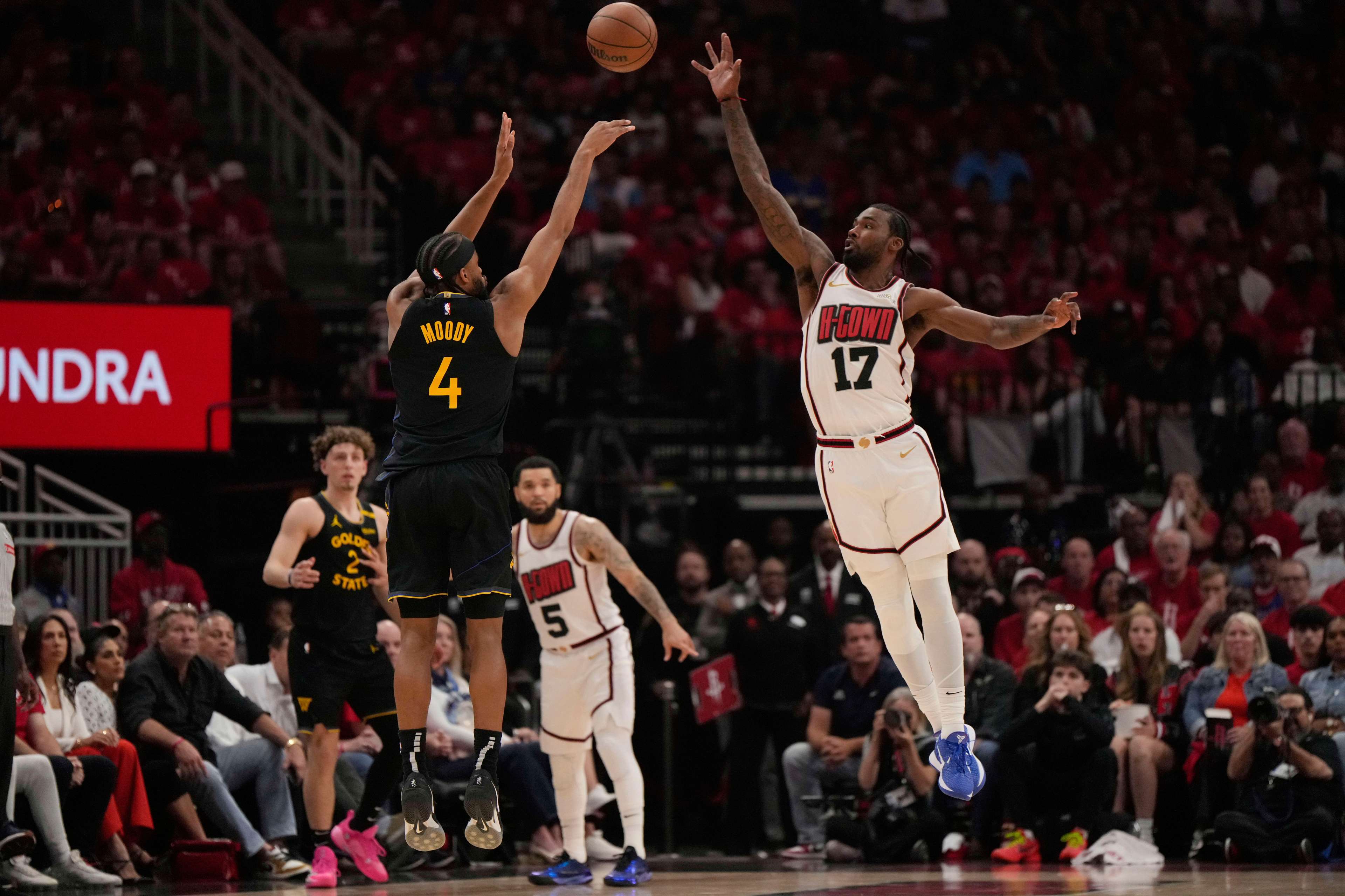 A basketball player wearing a black jersey numbered 4 is shooting, while another player in a white jersey numbered 17 jumps to block. Spectators watch intently.