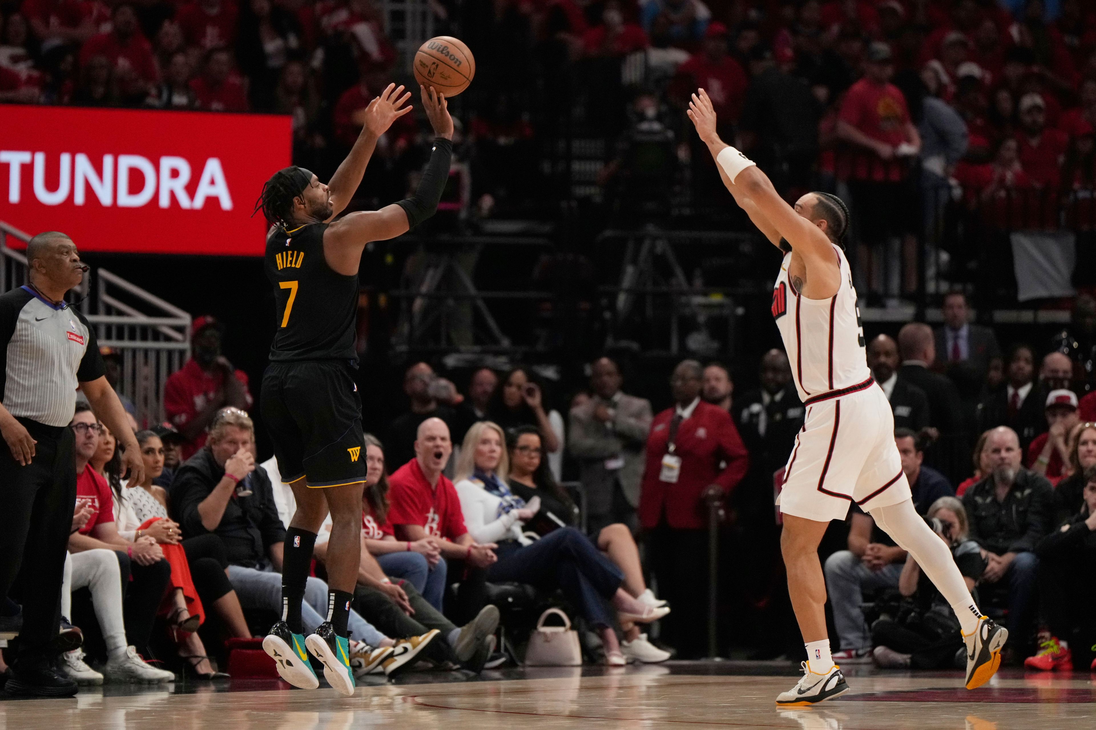 A basketball player in a black uniform jumps to shoot a ball, opposed by a player in a white uniform. A referee and spectators are visible in the background.