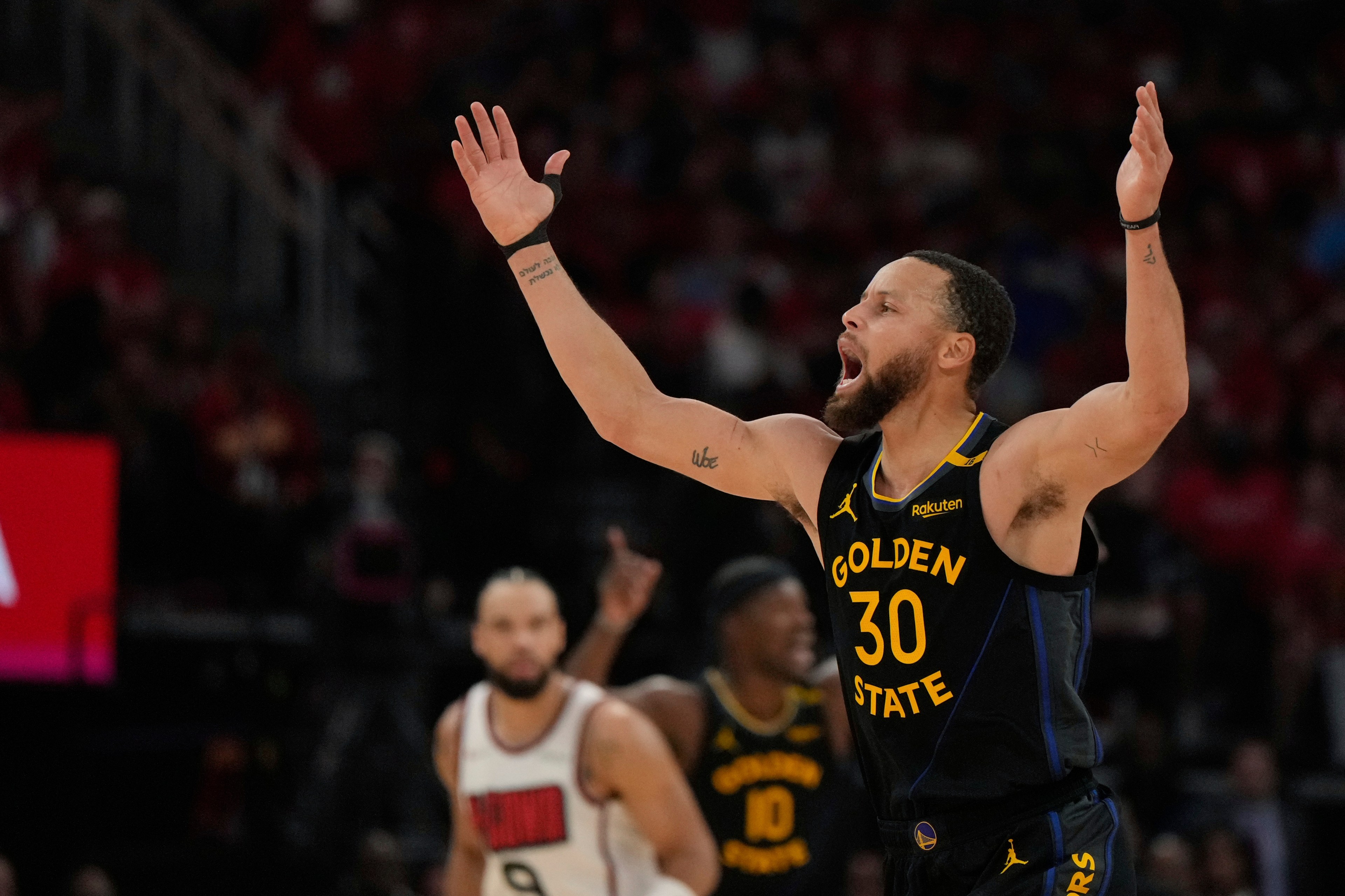 A basketball player wearing a Golden State jersey with the number 30 is raising his arms and shouting, while other players are visible in the background. The scene is intense.