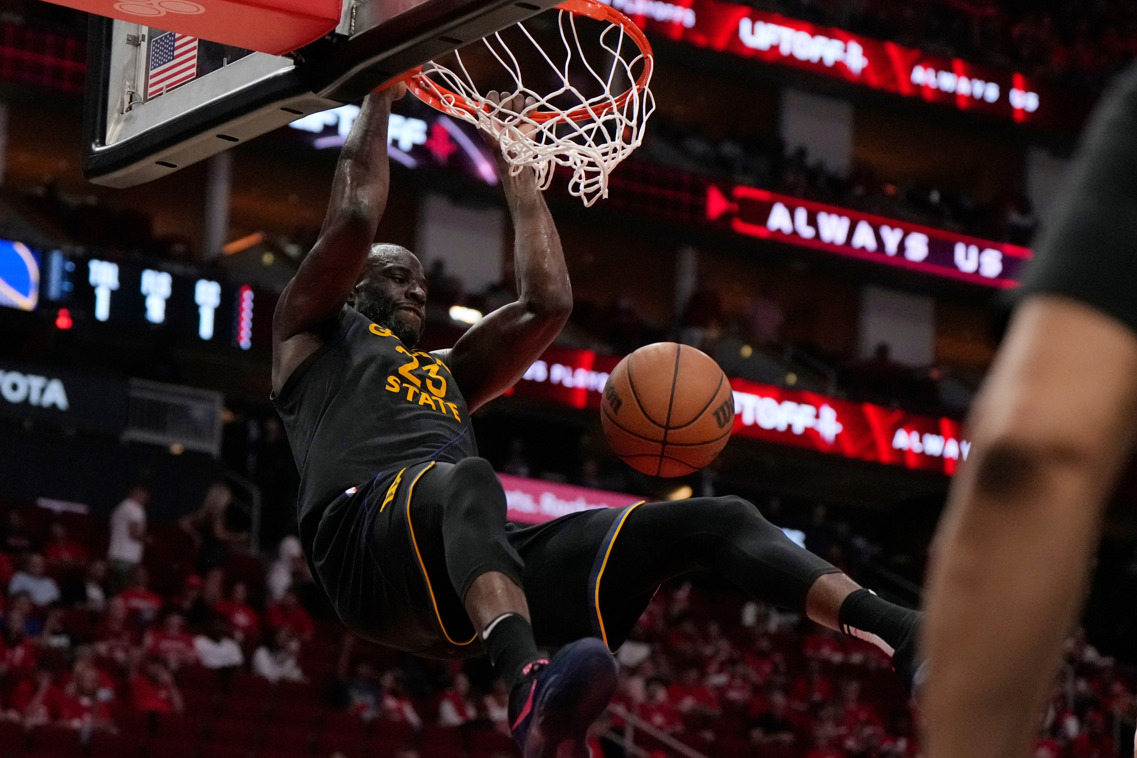 A basketball player in a black "Golden State" jersey hangs from the hoop after a slam dunk, with the ball dropping toward the floor in an indoor arena.
