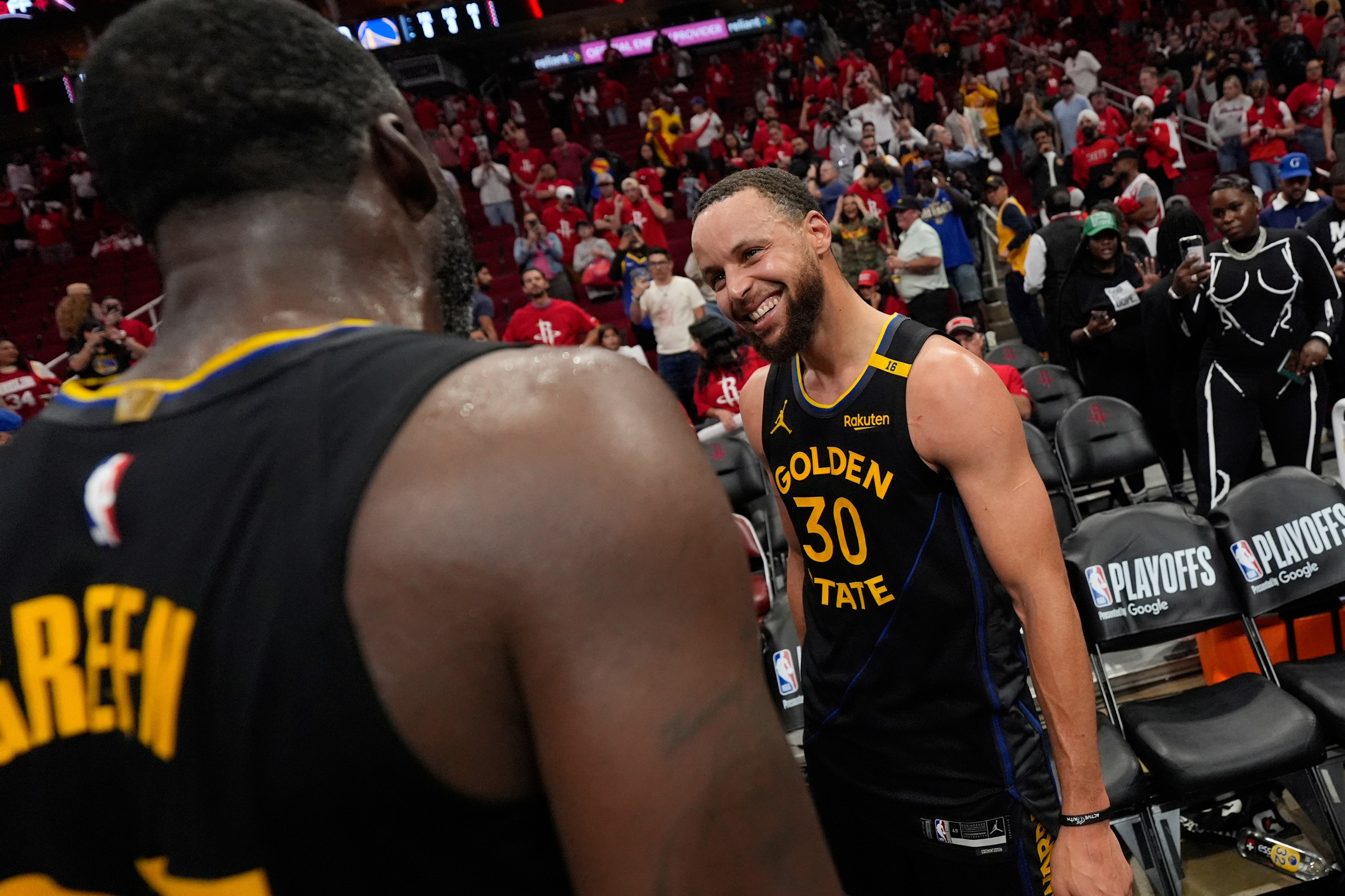 Two basketball players in Golden State jerseys share a moment on the court, smiling amid a crowd background, suggesting a post-game scene with fans.