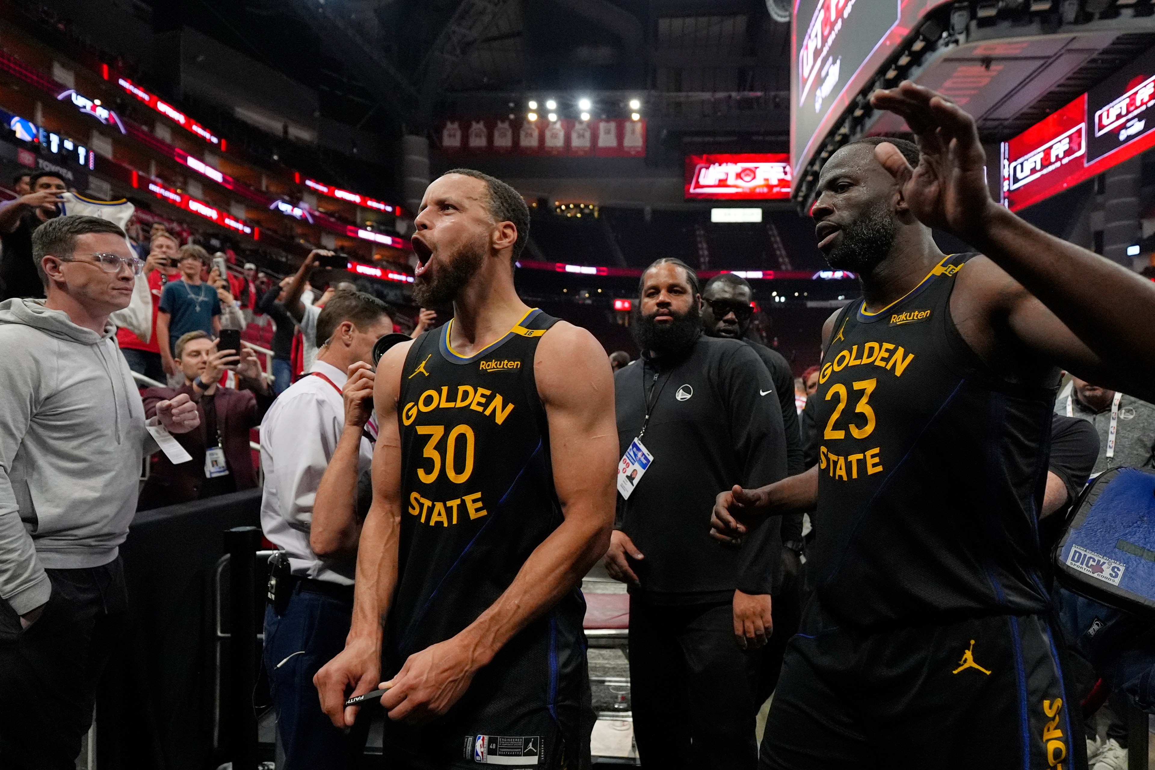 Two basketball players in Golden State jerseys, numbered 30 and 23, walk off the court, with one looking energized. Fans and staff surround them, taking photos.