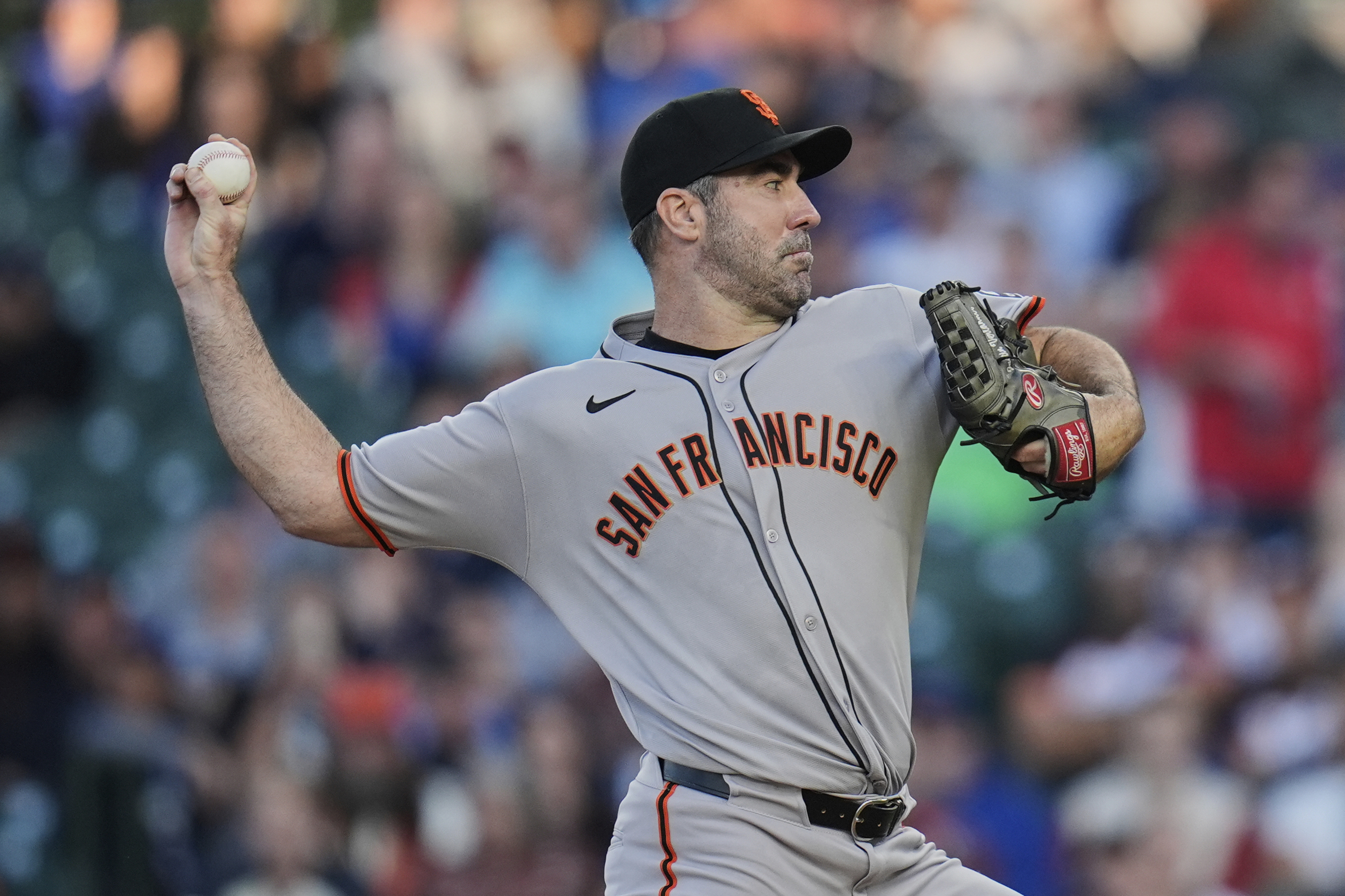 A baseball player is pitching, wearing a gray &quot;San Francisco&quot; jersey, a black cap, and a glove. The blurred crowd is in the background.