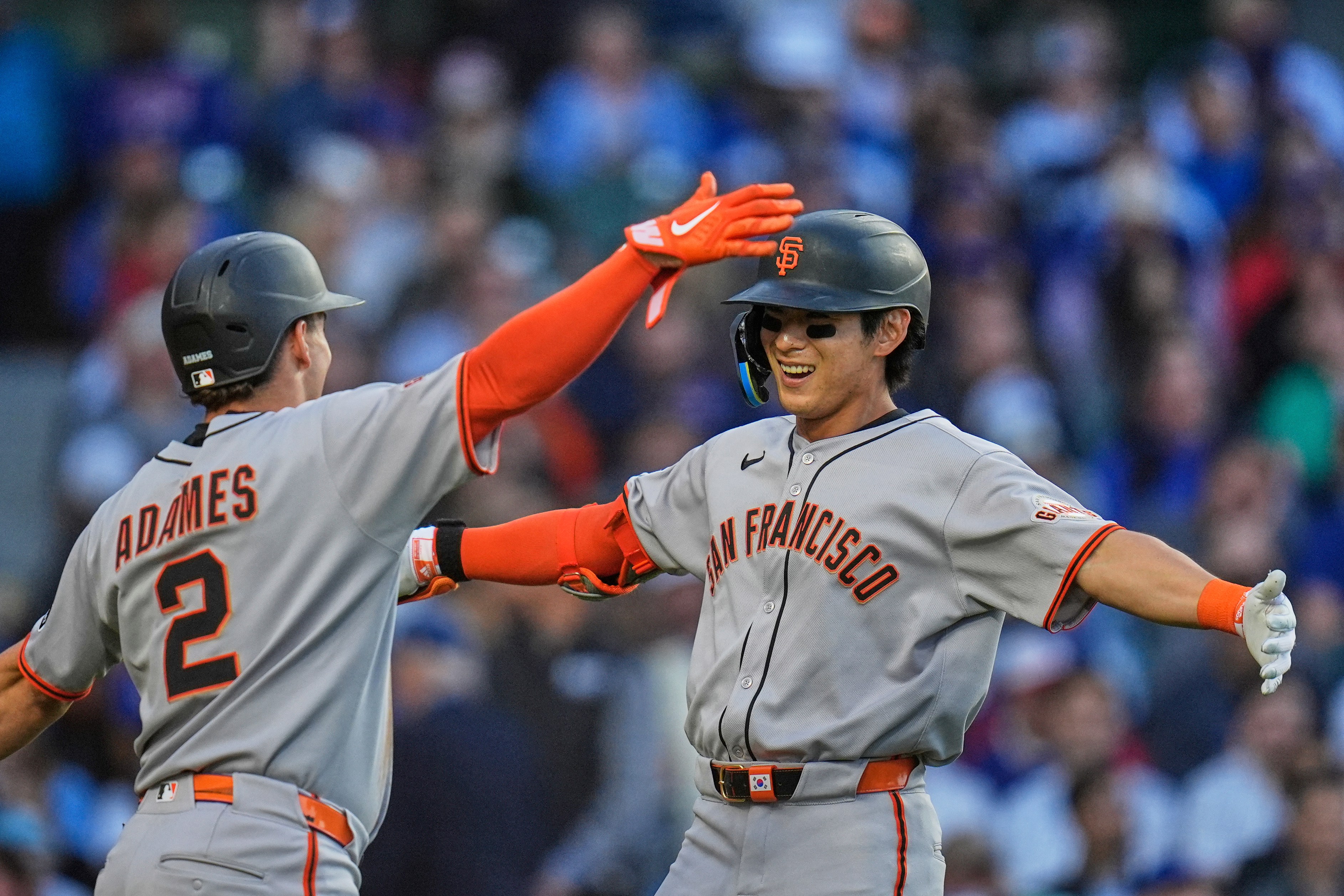 Two baseball players in San Francisco uniforms celebrate on the field, doing a high-five. One wears a helmet labeled "ADAMES." Both appear joyful.