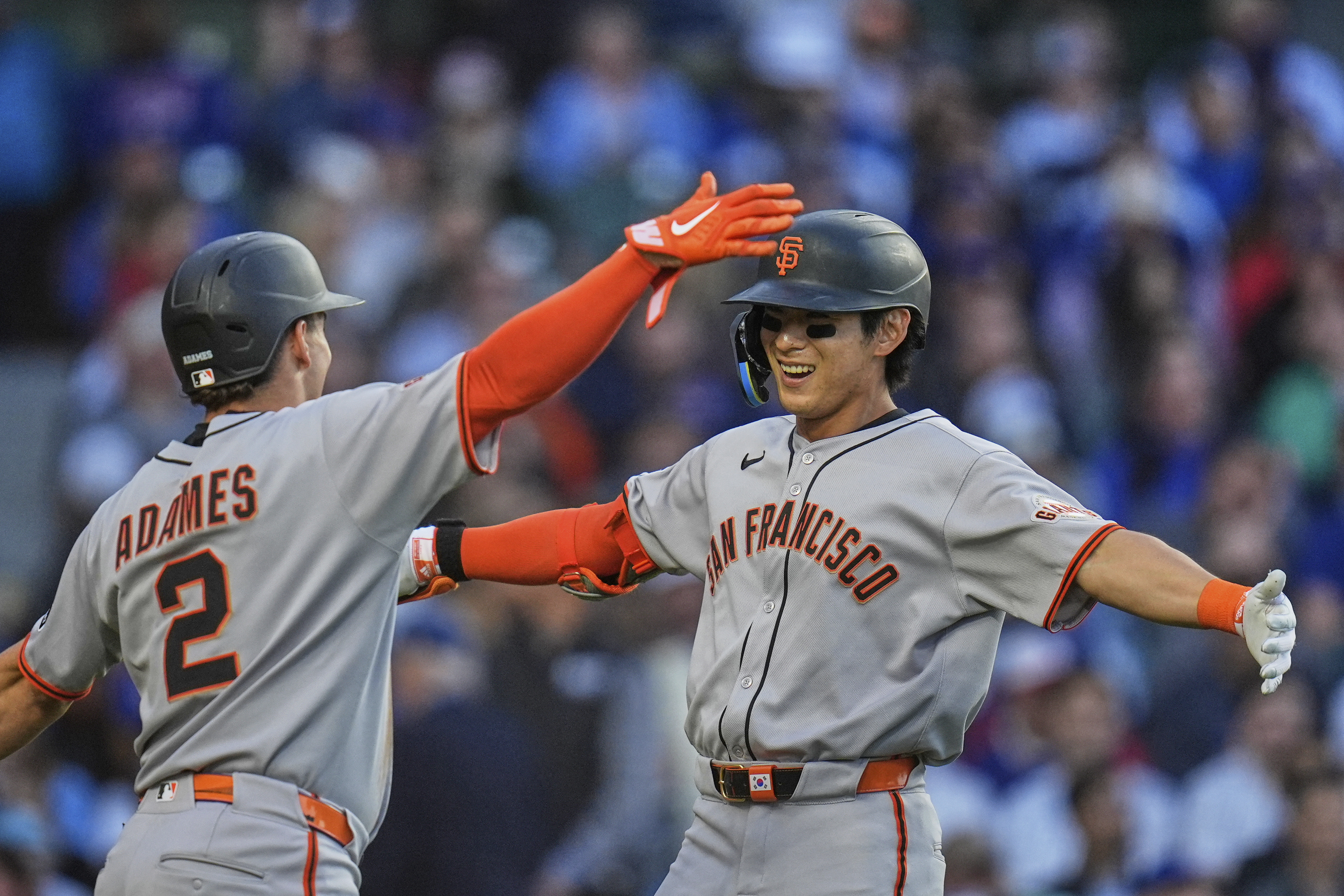 Two baseball players in San Francisco uniforms celebrate on the field, doing a high-five. One wears a helmet labeled "ADAMES." Both appear joyful.
