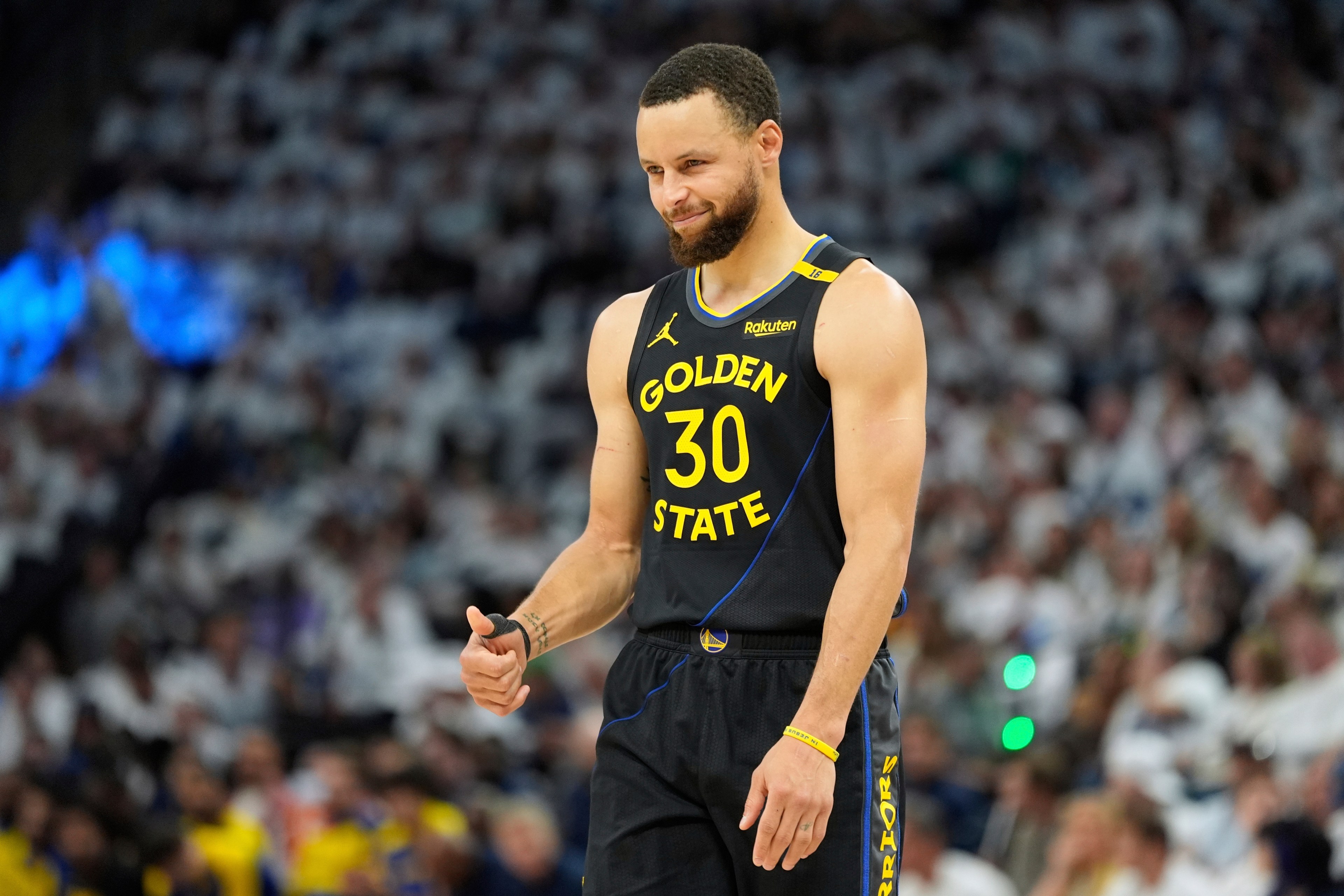 A basketball player in a black Golden State jersey (#30) gives a thumbs-up. He smiles slightly, standing on the court with a blurred crowd in the background.