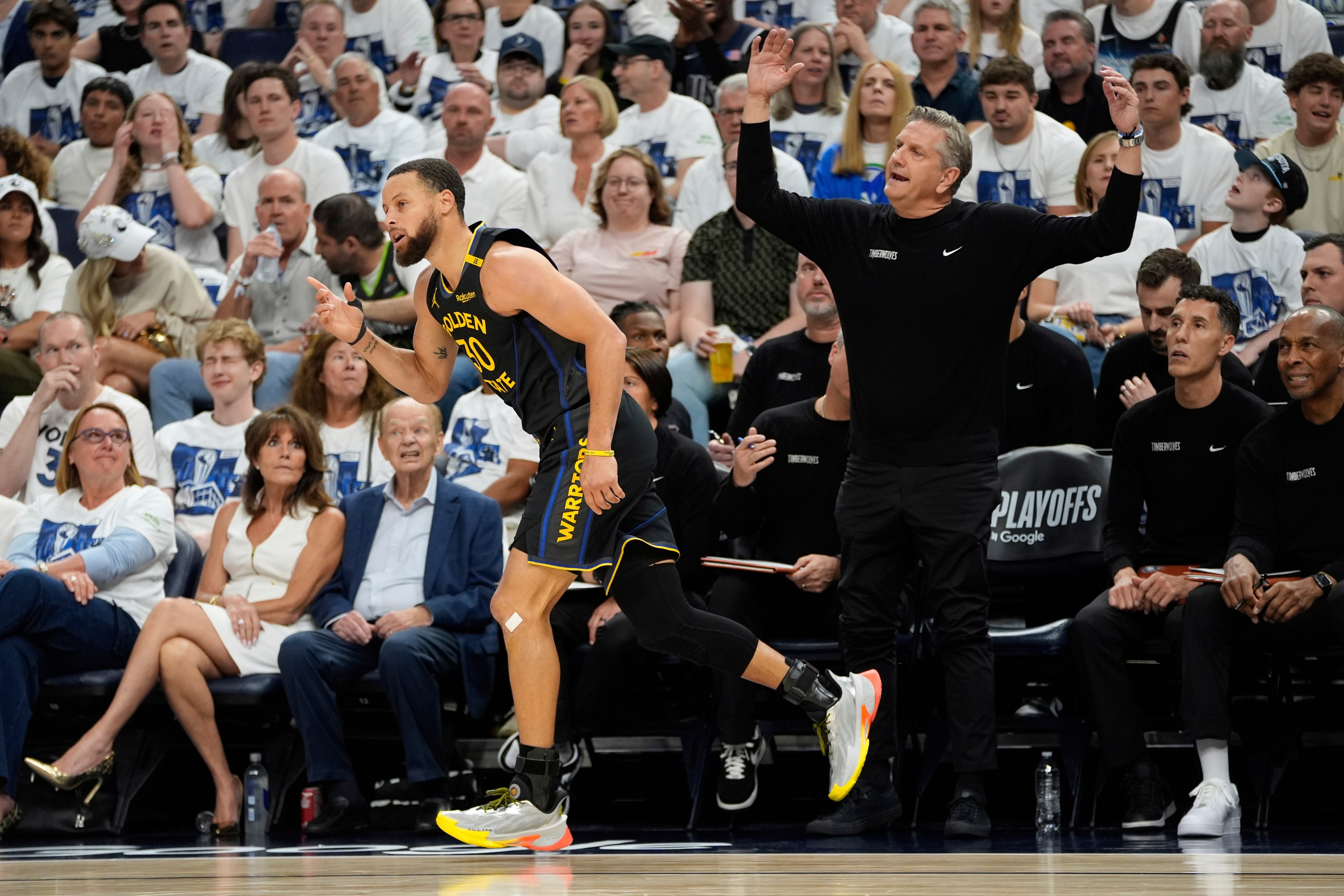 A basketball player in a Warriors uniform is running on the court. Nearby, a coach on the sideline gestures, while fans in white shirts watch intently.