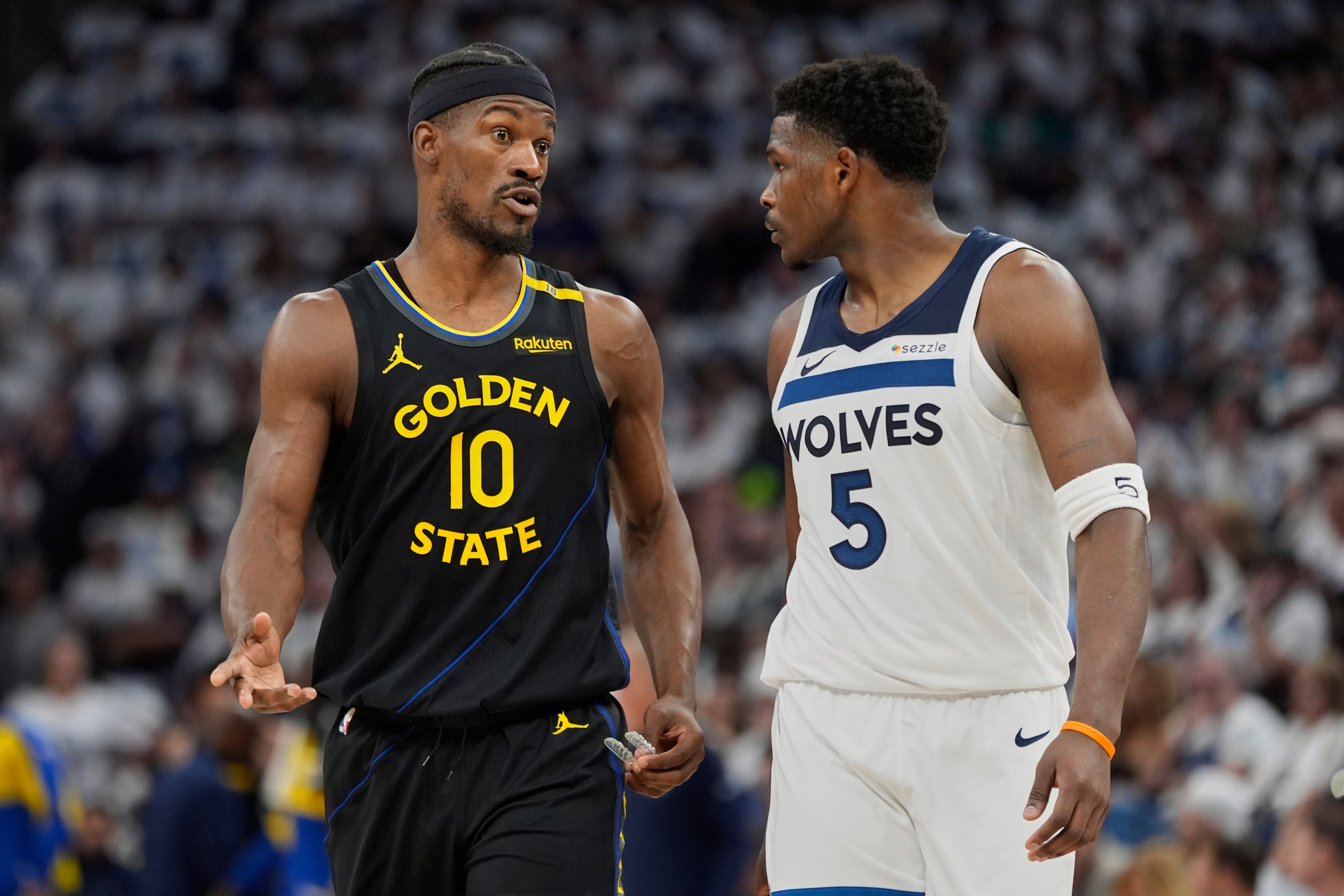Two basketball players are on the court. One wears a black Golden State jersey, the other a white Wolves jersey. They're talking during a game.