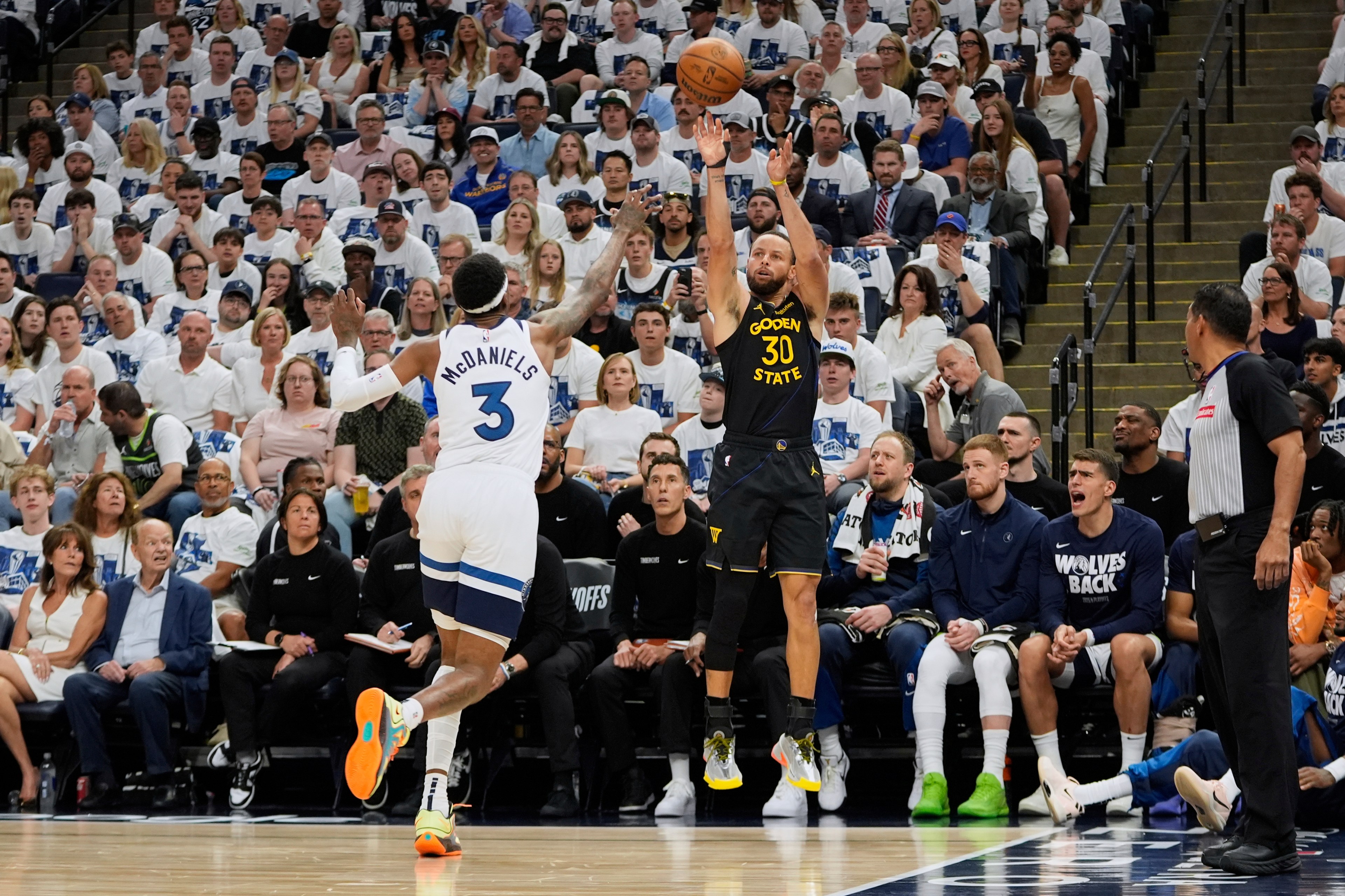 A basketball player in a black &quot;Golden State&quot; jersey is shooting a jump shot over a defender in a white jersey with &quot;McDaniels&quot; on it. The crowd is attentively watching.