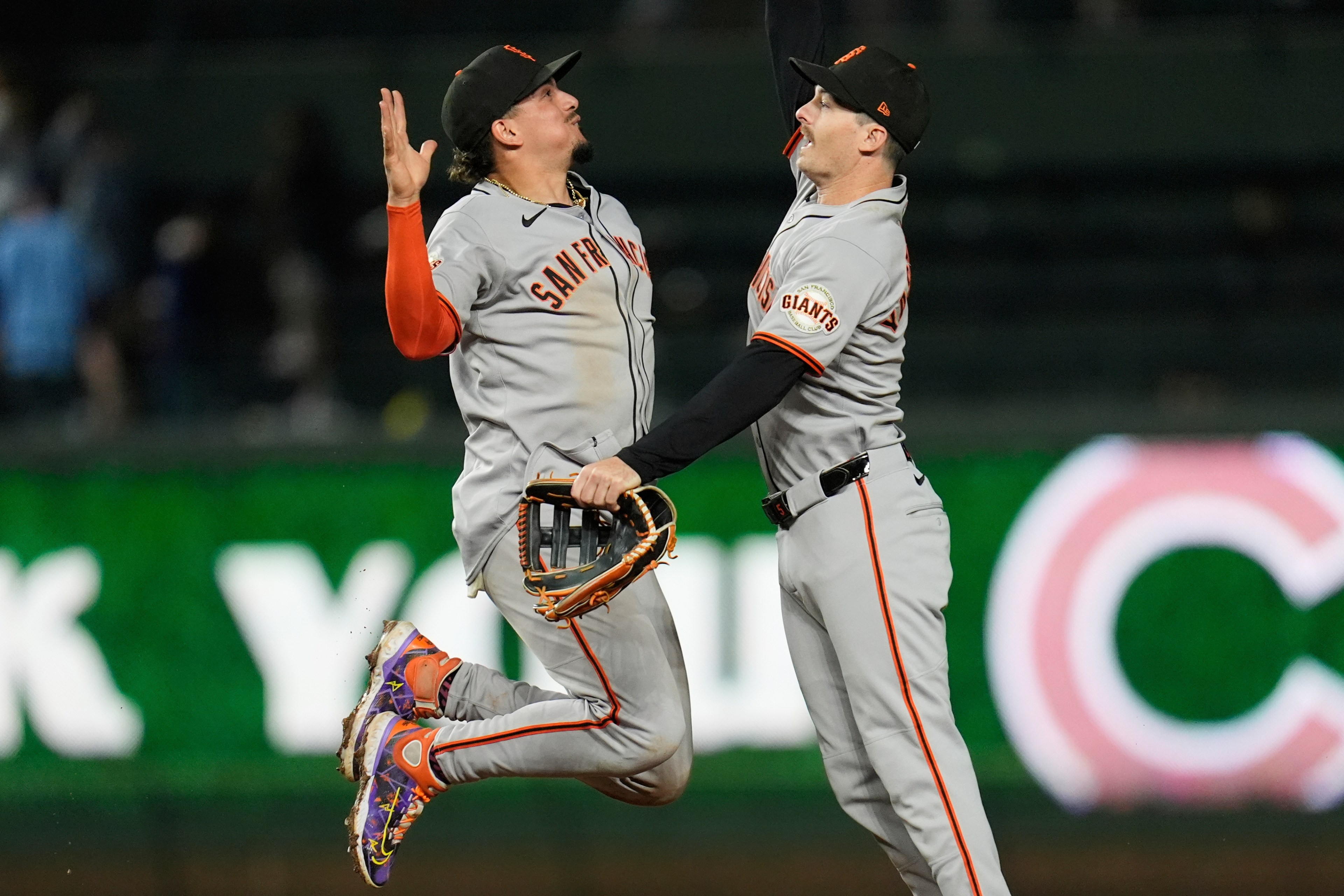 Two baseball players in gray uniforms jump and high-five in celebration on the field. One player holds a glove, and both are wearing black caps with orange details.