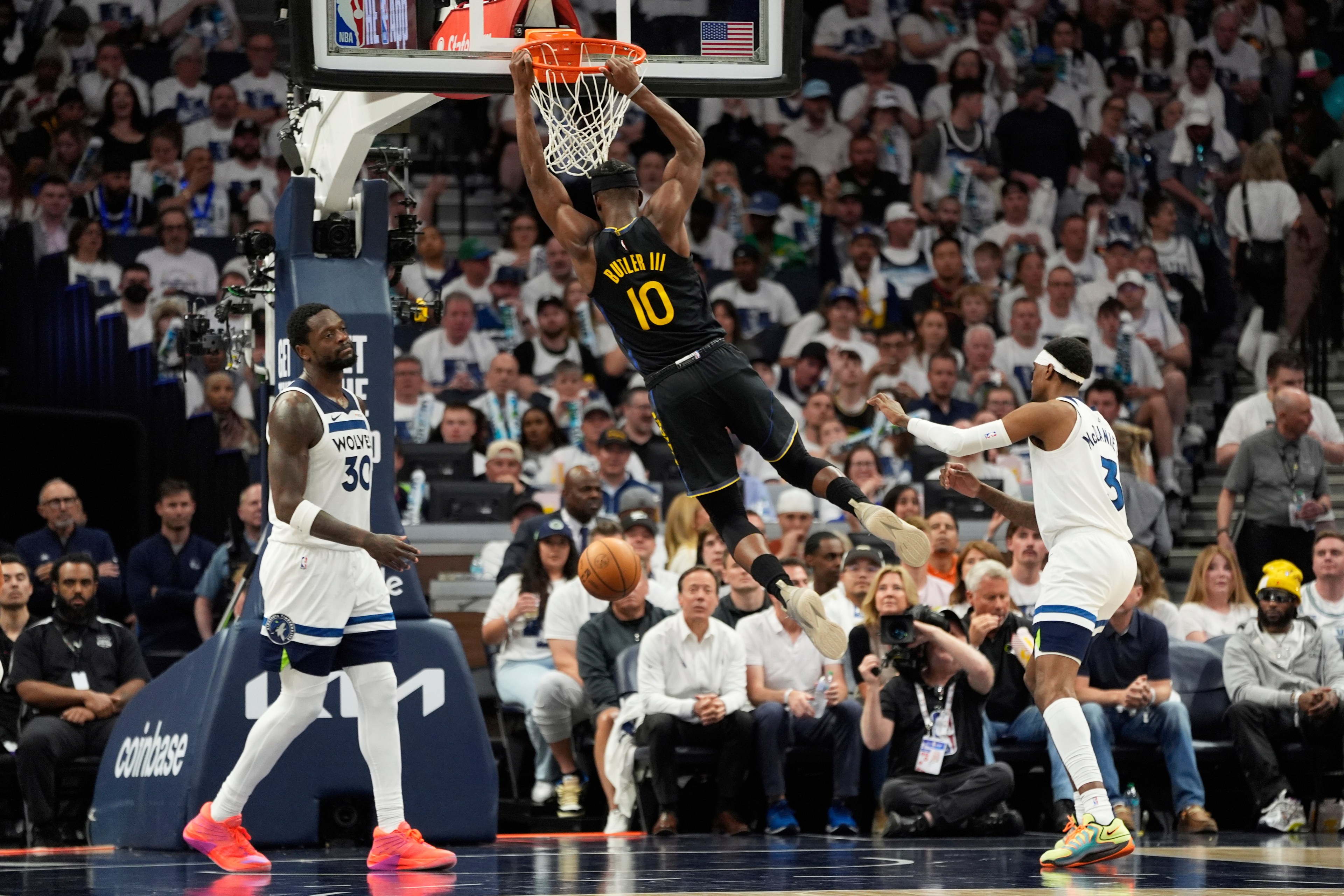 A basketball player in a black jersey dunks the ball while two players in white jerseys watch. The crowd in the background appears engaged.