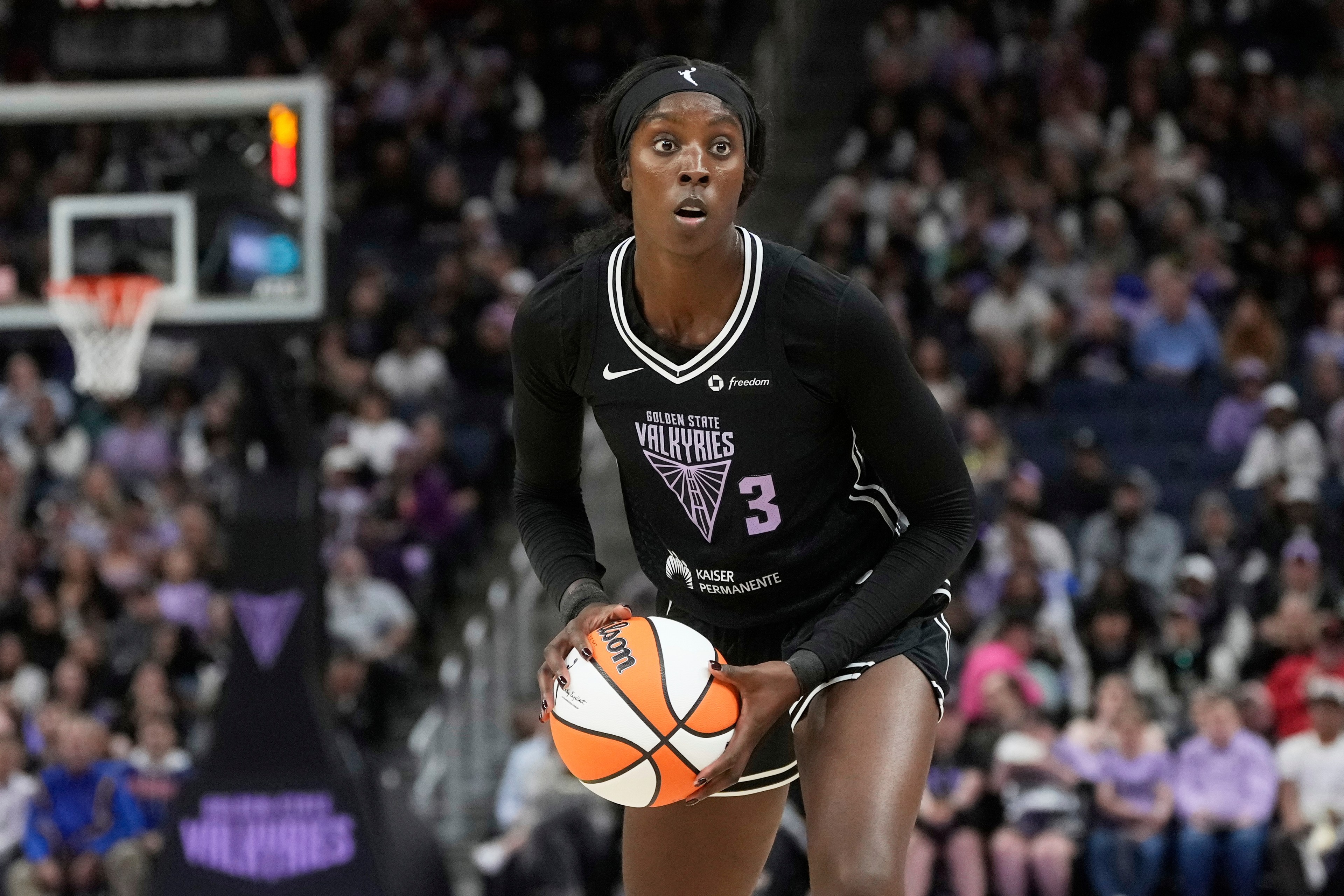 A basketball player in a black jersey is holding a basketball in a court. The crowd is blurred in the background, and the jersey reads &quot;Golden State Valkyries.&quot;