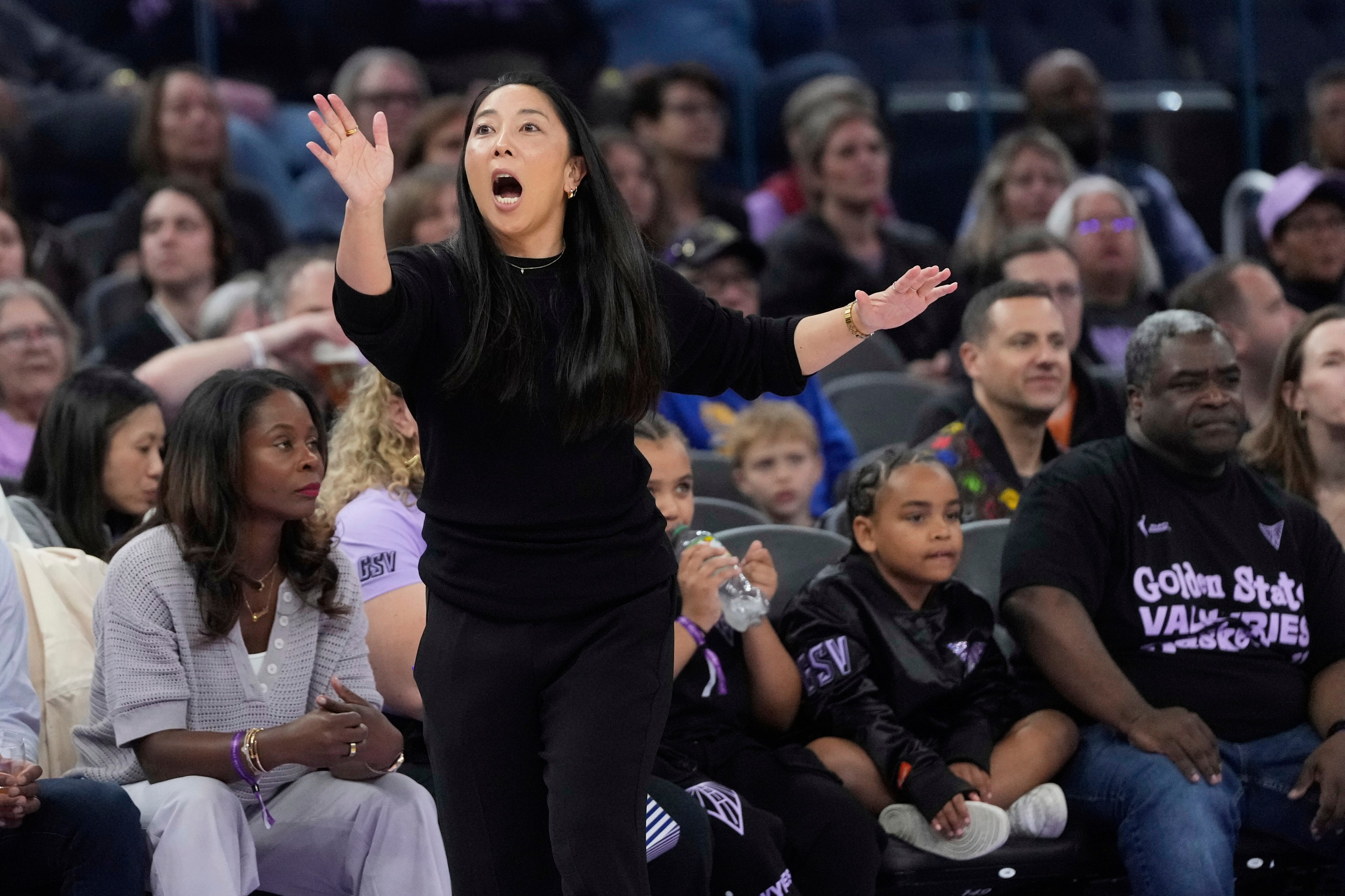 A woman in black passionately gestures from the sidelines at a sports event. Spectators fill the background, some focused on the action.