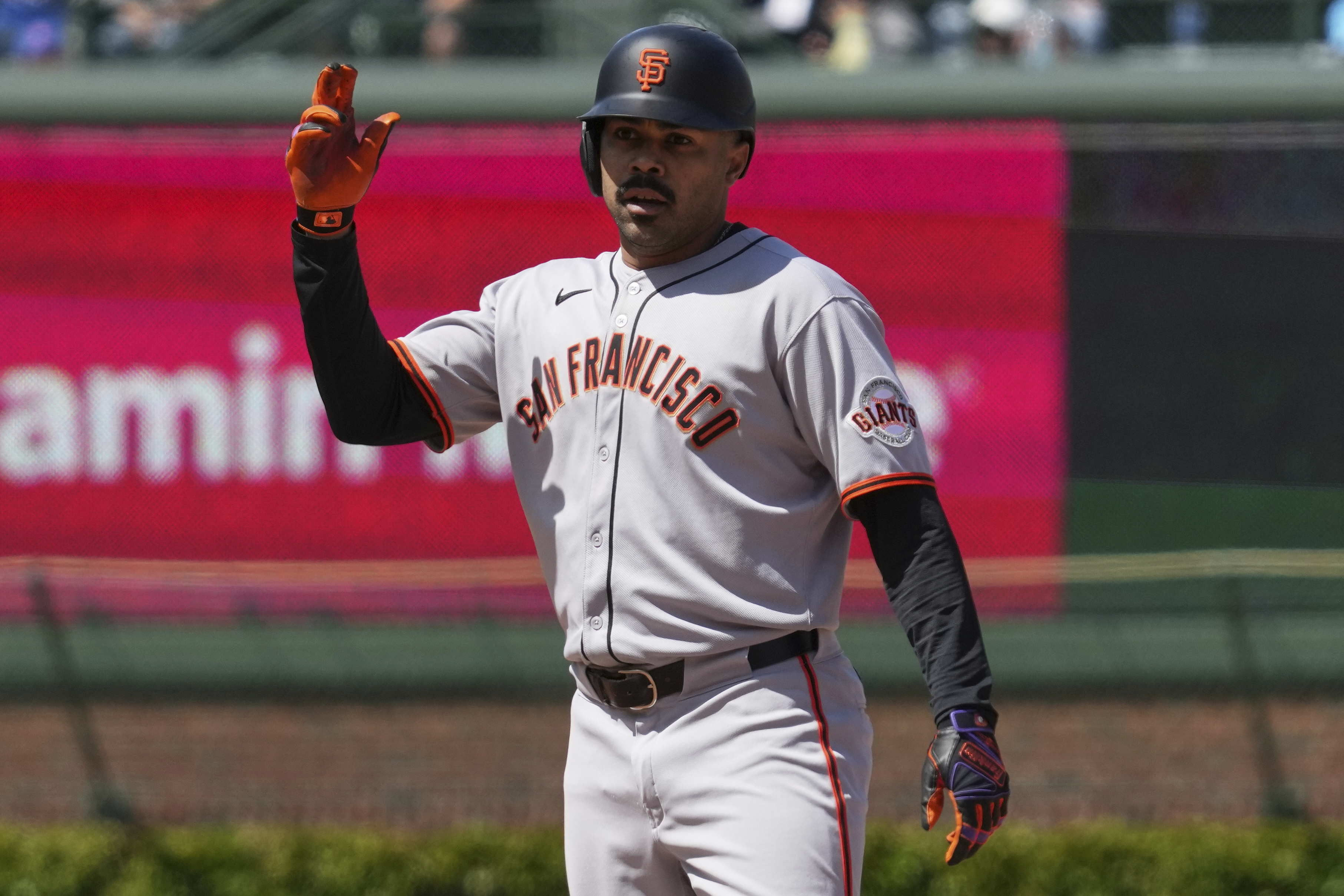 A baseball player wearing a San Francisco Giants uniform and helmet holds up his left hand. He's on a field with blurred banners in the background.