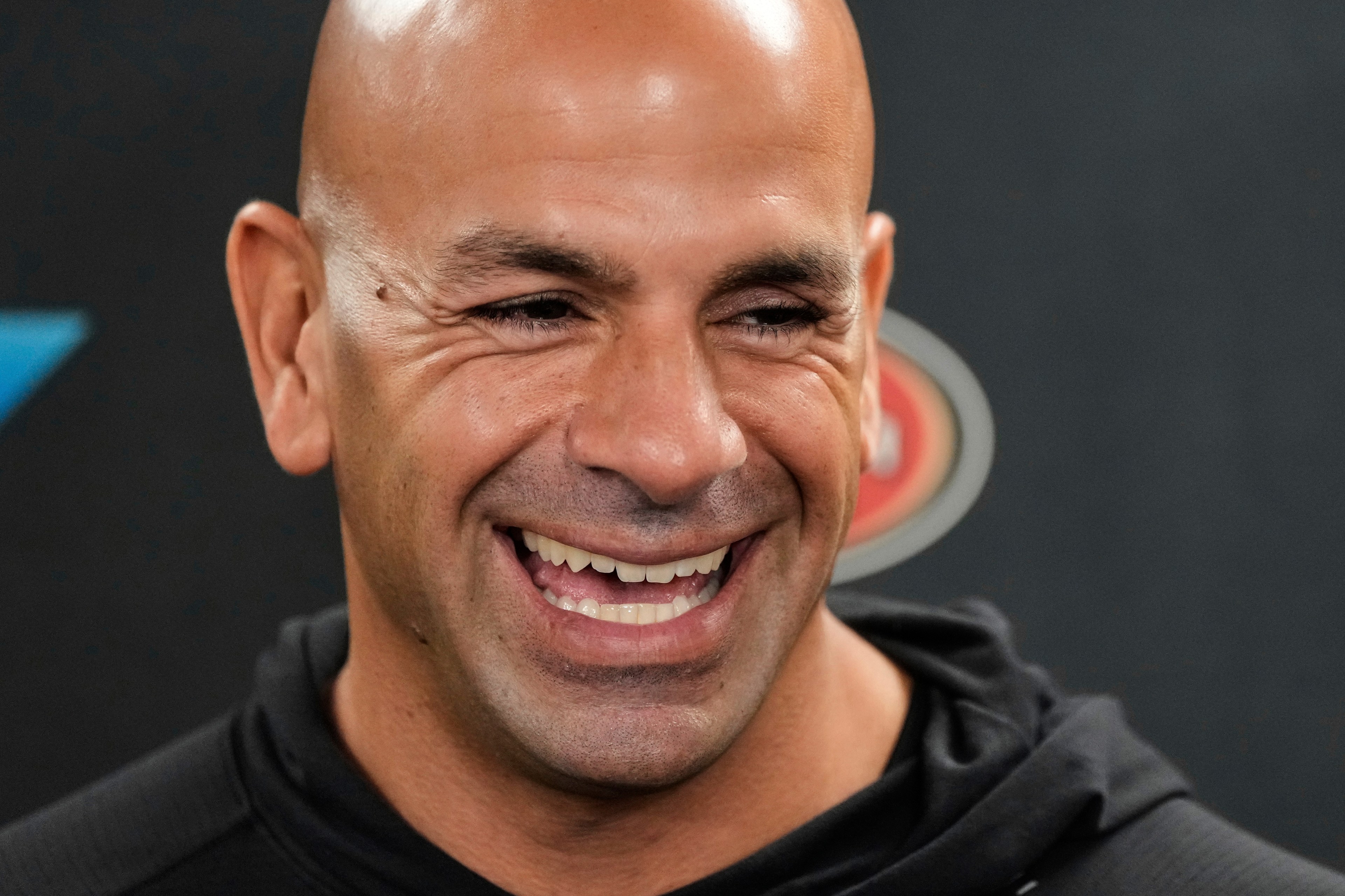 A man with a bald head and a broad smile is wearing a black hoodie. He has deep laugh lines, and there's a blurred logo in the background.