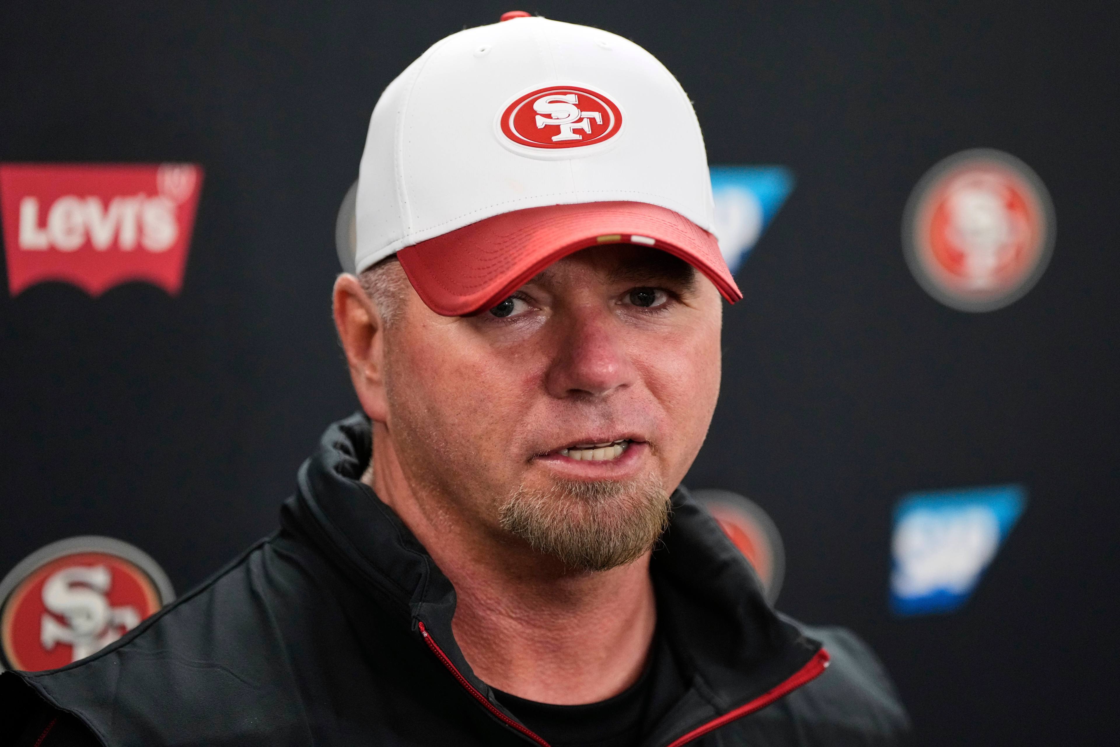 A man in a white and red cap with a &quot;49ers&quot; logo is speaking at a press event. The background features &quot;Levi's&quot; and &quot;SAP&quot; logos.