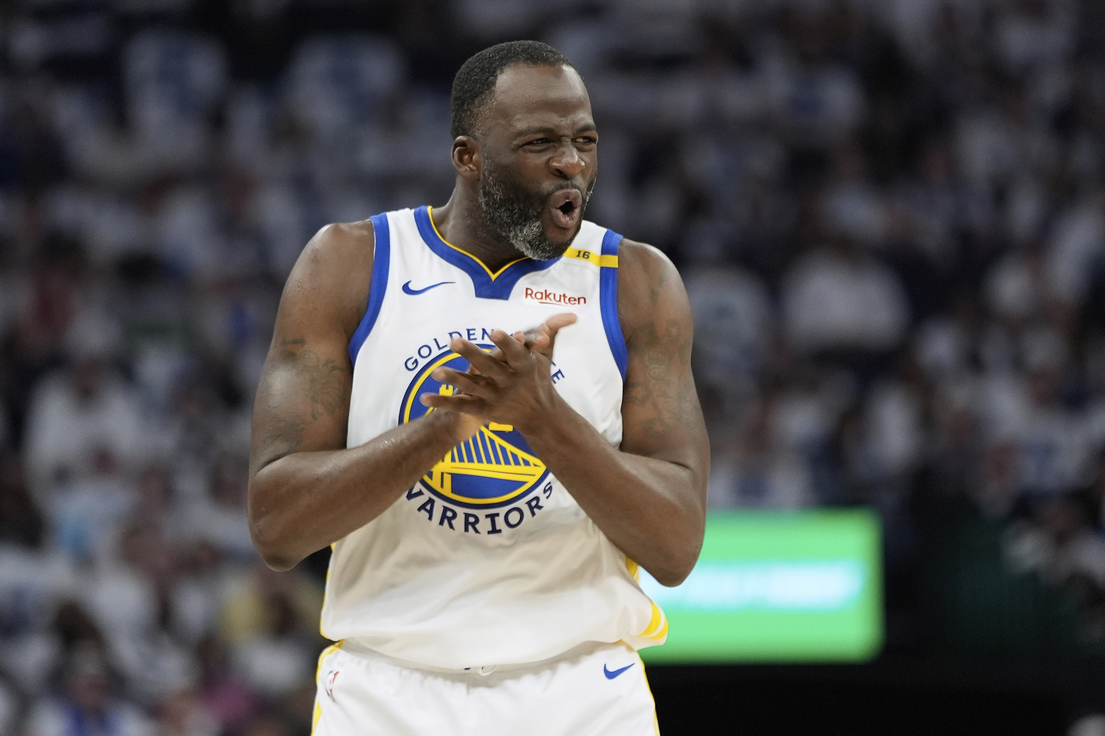 A basketball player in a white Golden State Warriors jersey claps his hands, showing an expressive facial expression, with a blurred crowd in the background.