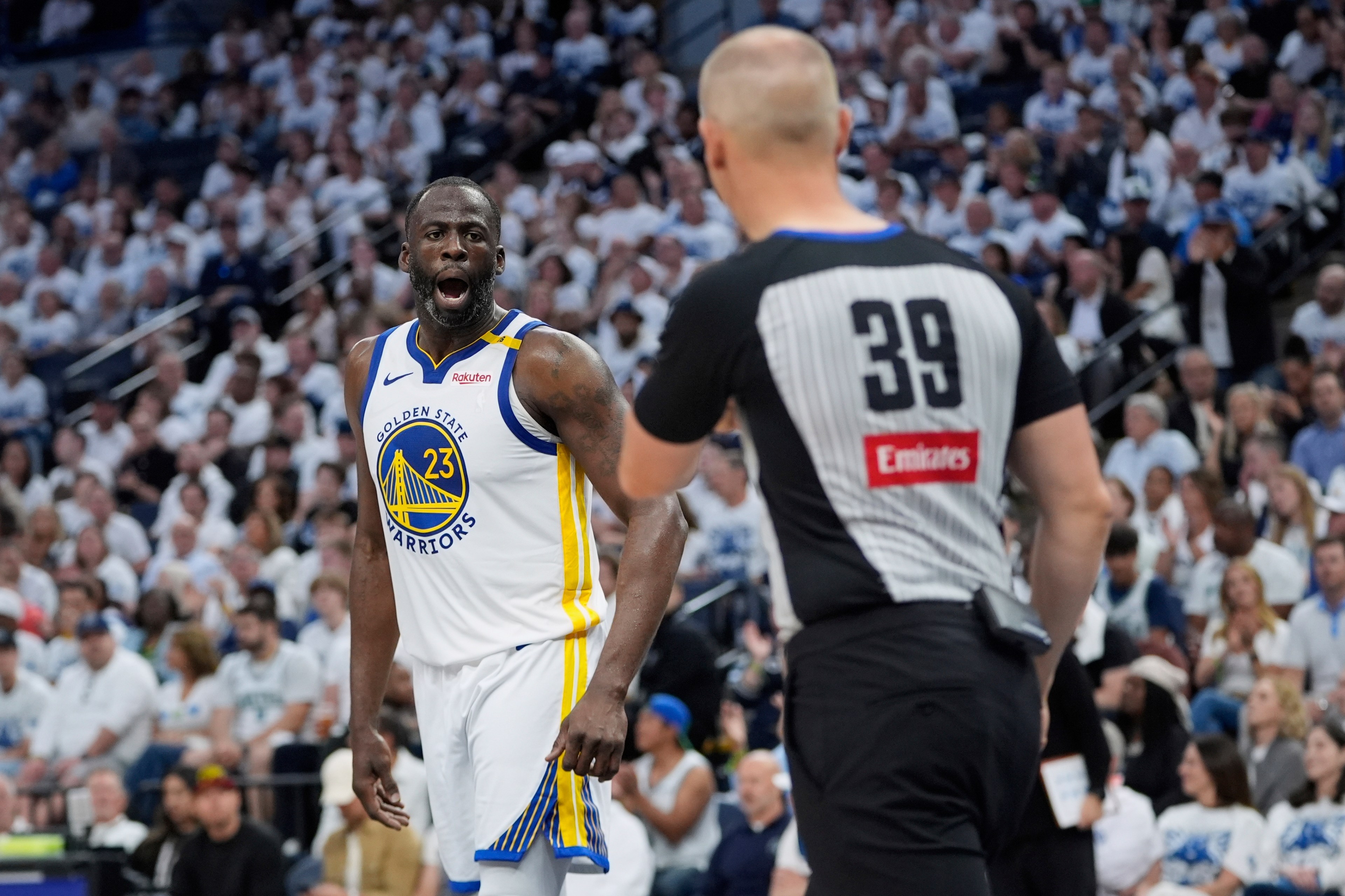 A basketball player in a white Golden State Warriors jersey is animatedly talking to a referee in a black-and-white striped shirt with "39" on the back.