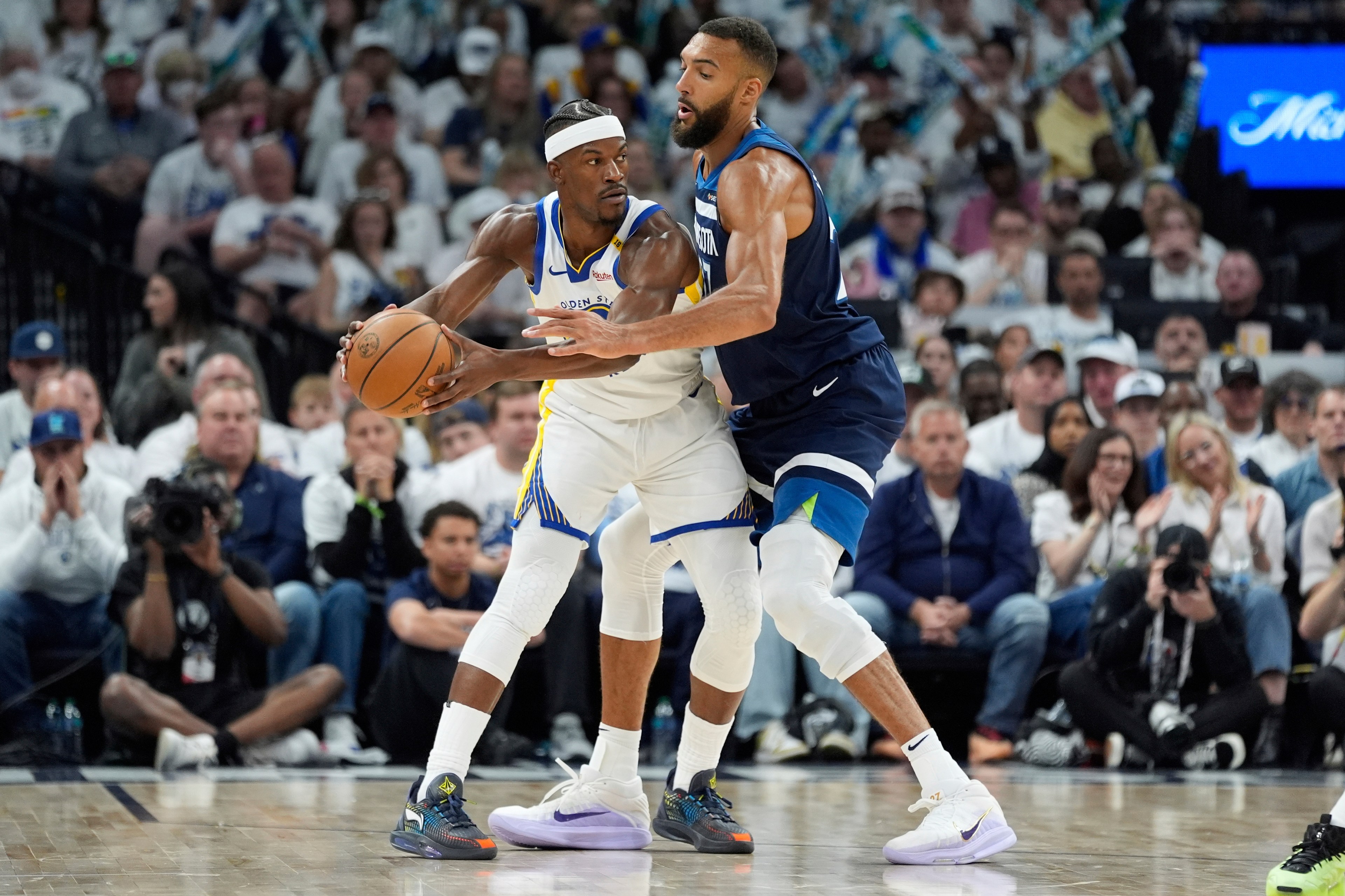 Two basketball players are on a court, one in a white jersey holding the ball, and the other in a dark jersey defending. A crowd watches intently.