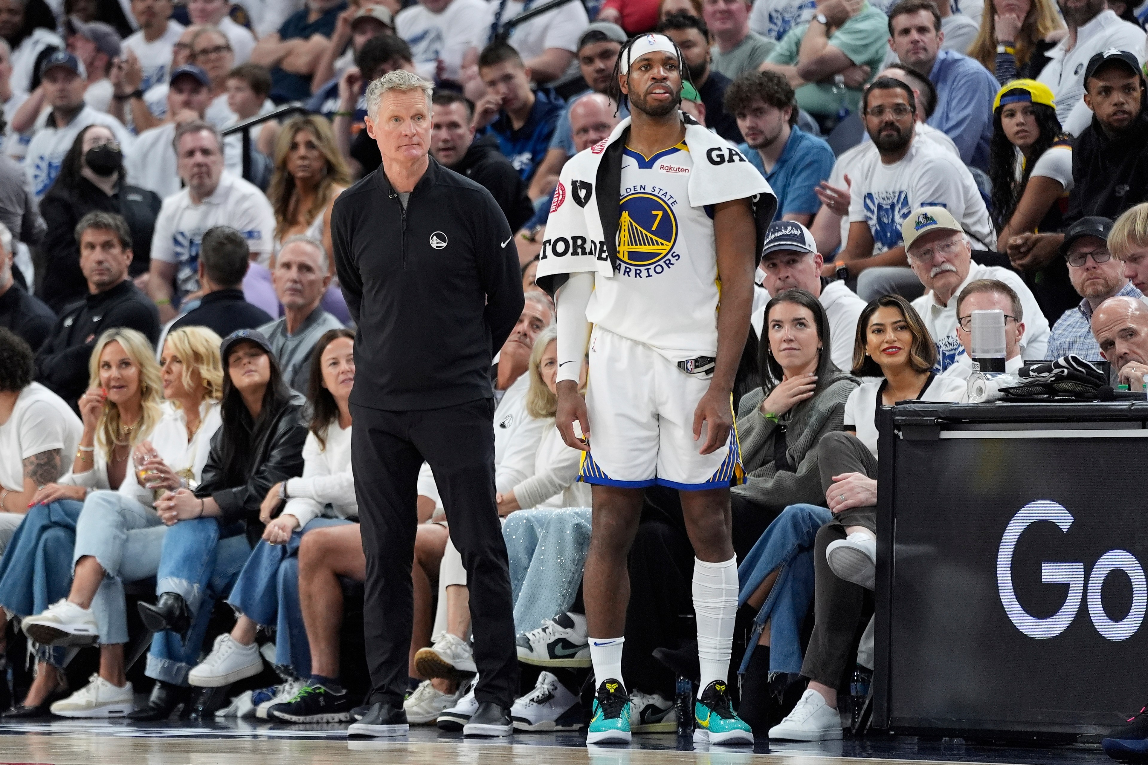 A basketball coach stands beside a player in a Golden State Warriors jersey with a towel draped over his shoulder. They're in front of a crowded, attentive audience.