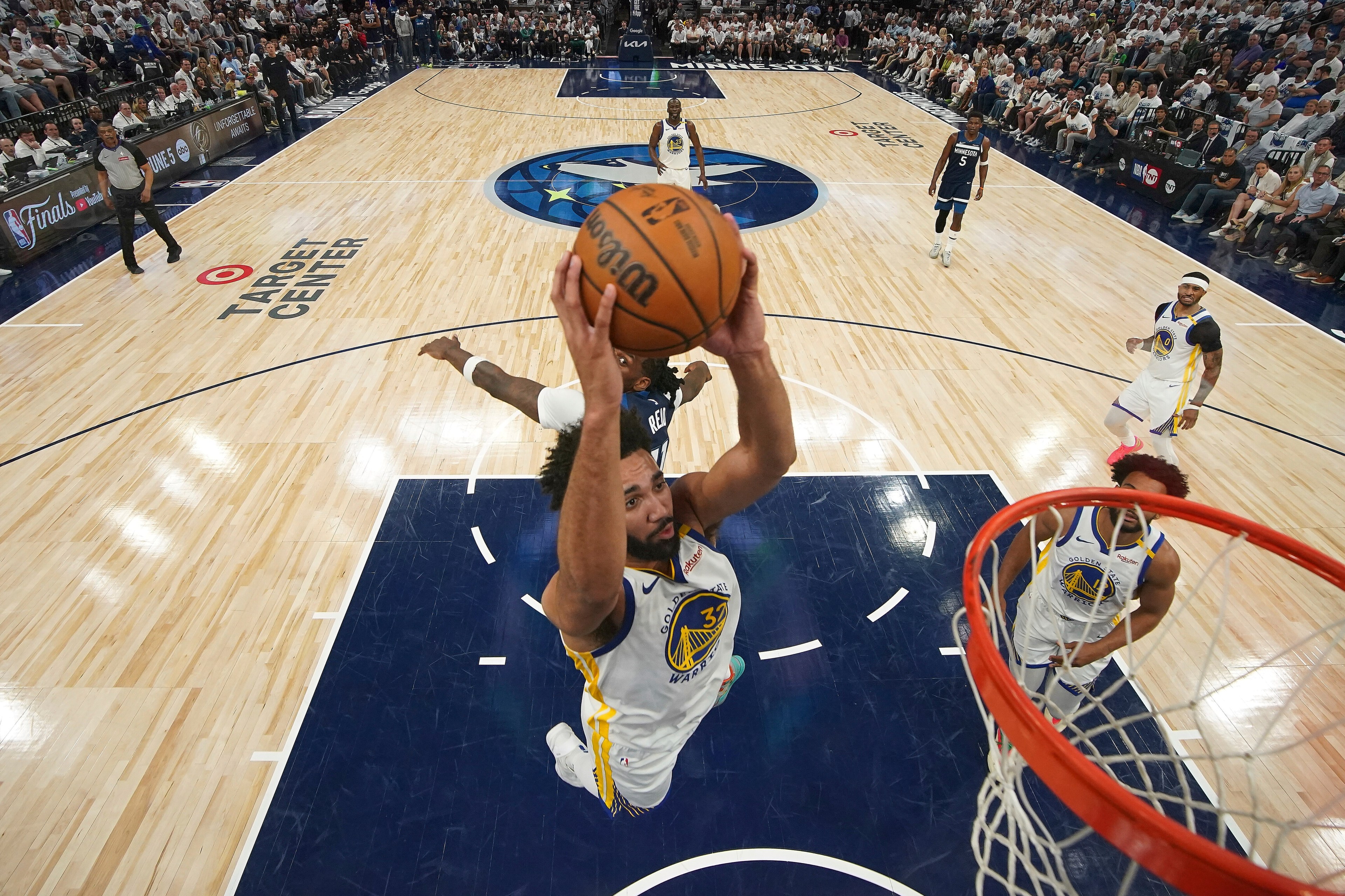 A basketball player in a Warriors jersey jumps to score a basket, while a Timberwolves player attempts to block him on a crowded court at Target Center.