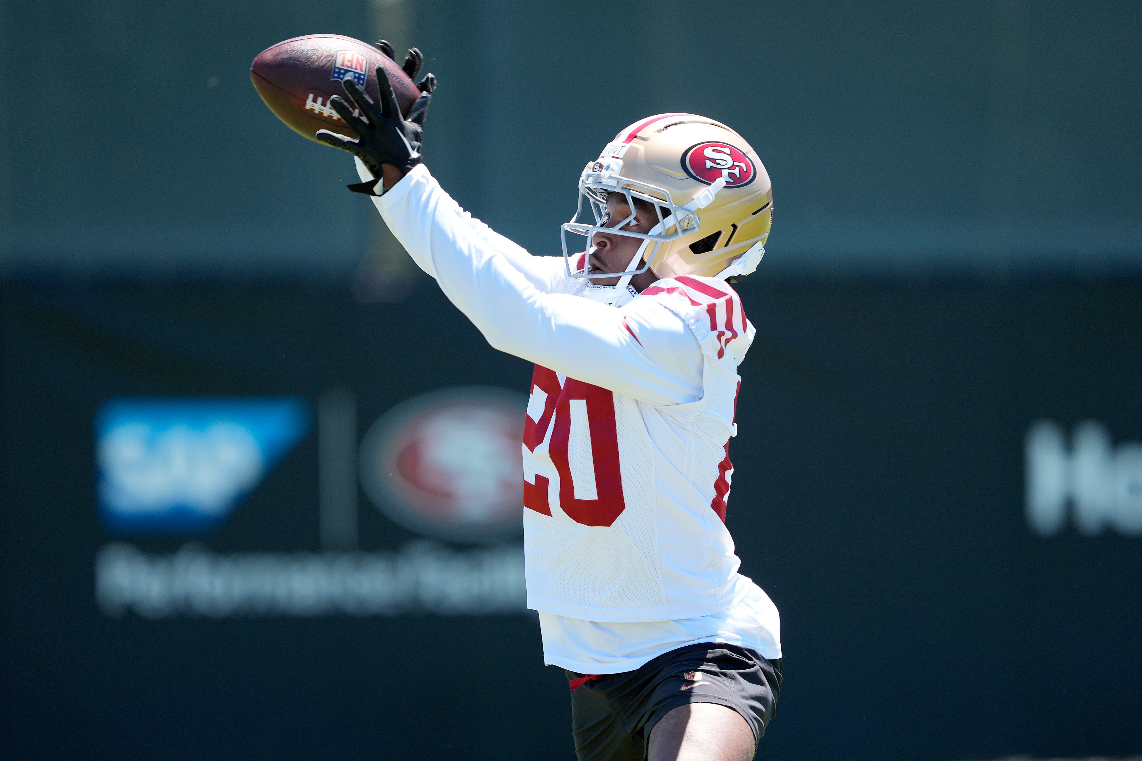 A football player in a red and gold helmet and white jersey is catching a football with both hands during practice, with a blurred background.
