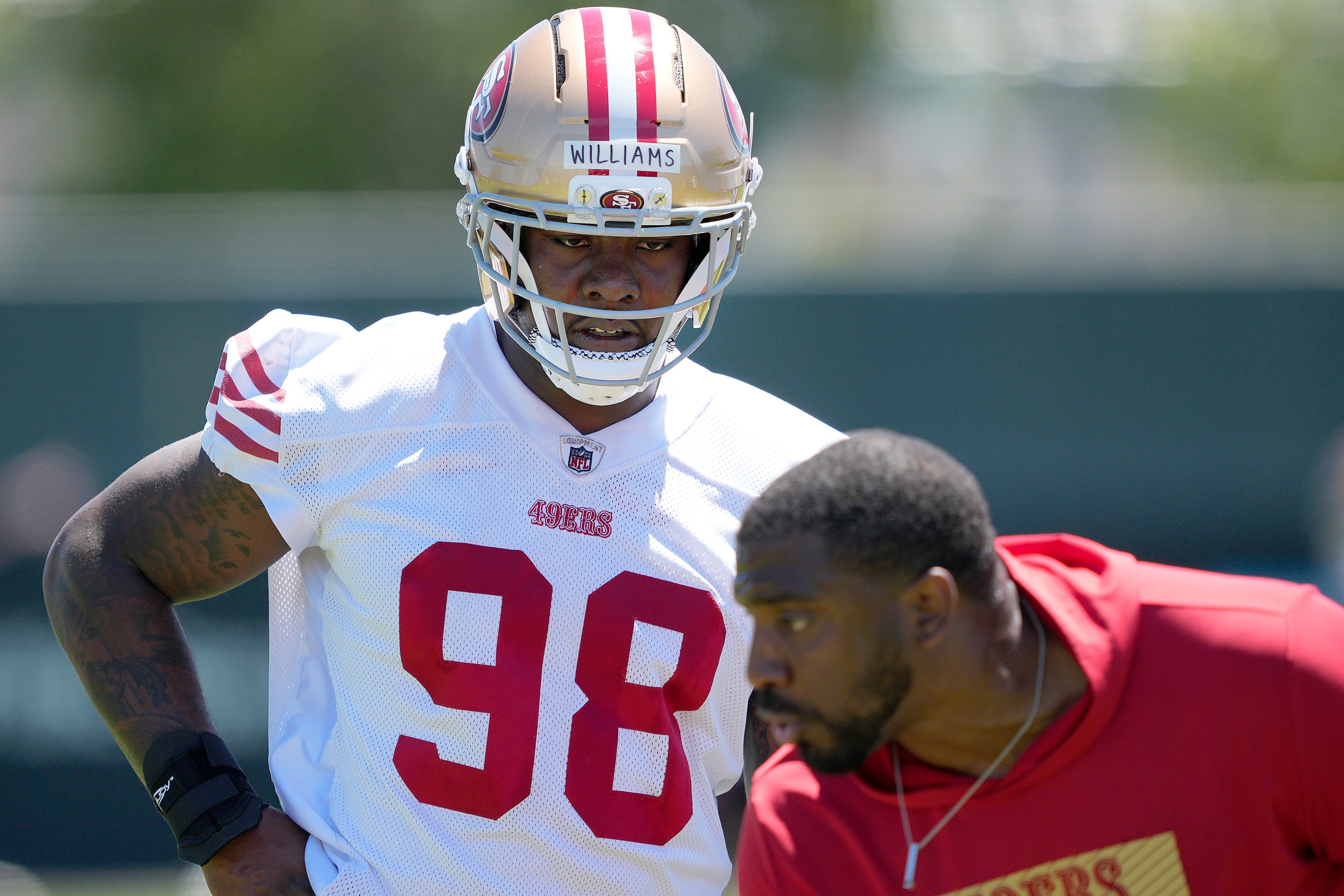 A football player wearing a 49ers uniform with number 98 practices on a field, accompanied by a coach in a red 49ers shirt.