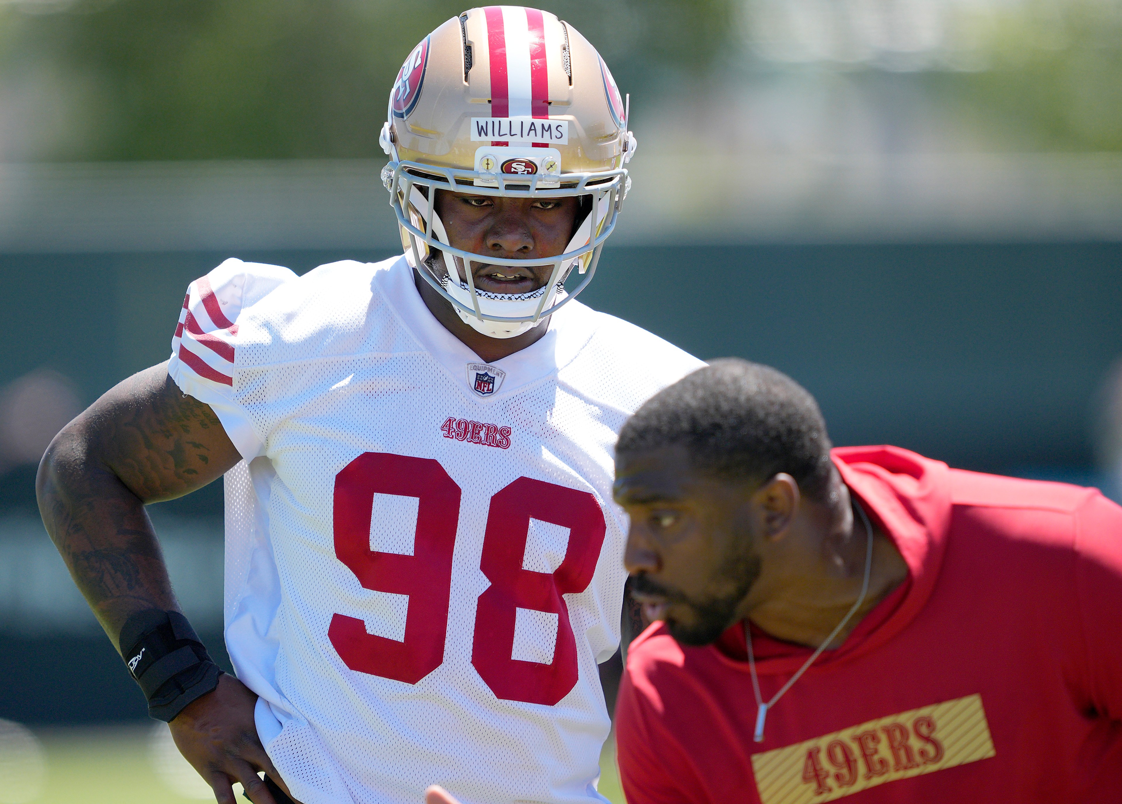 A football player wearing a 49ers uniform with number 98 practices on a field, accompanied by a coach in a red 49ers shirt.