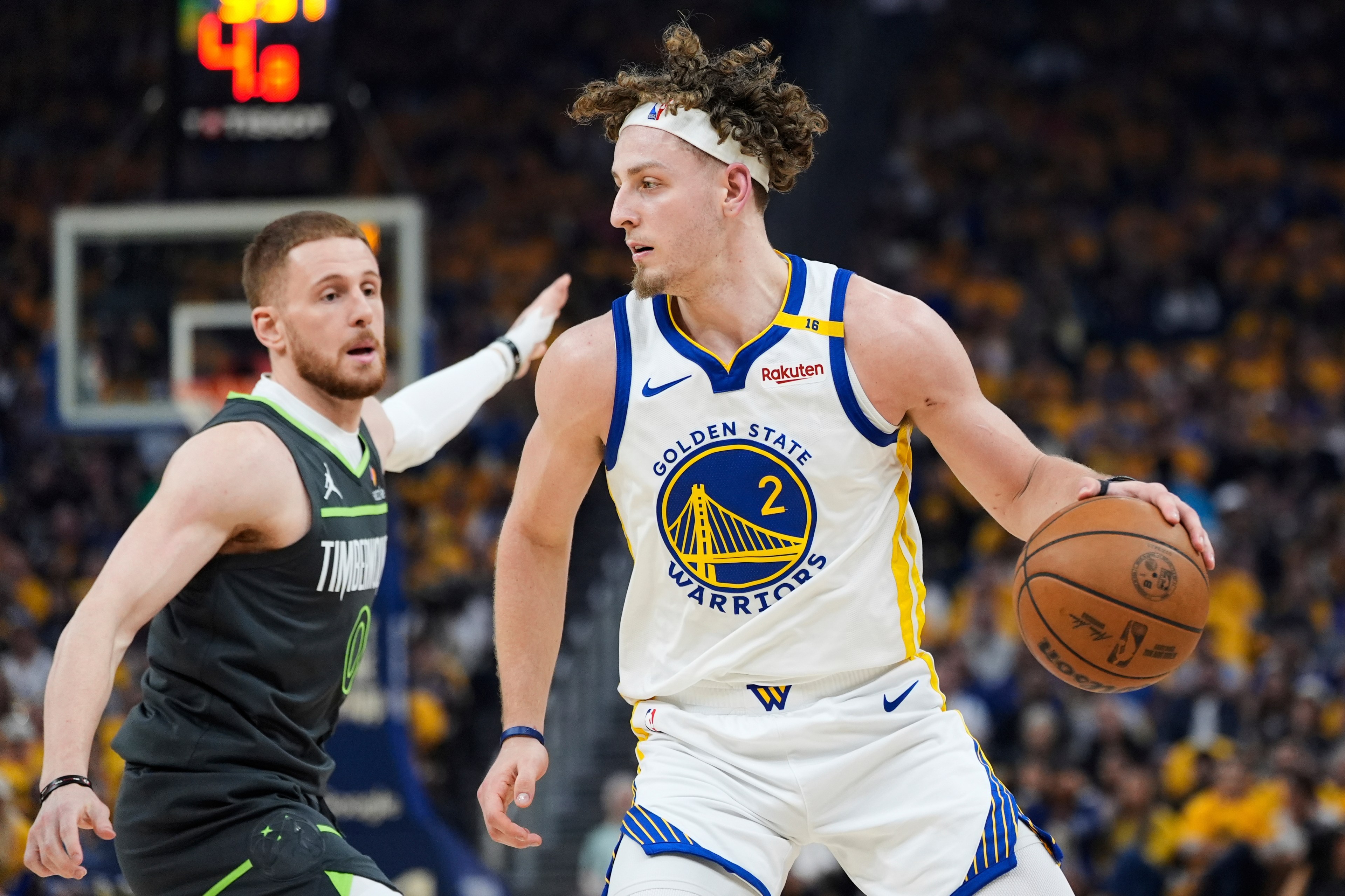 Two basketball players are on the court: one in a Golden State Warriors jersey dribbling the ball, and the other in a Timberwolves jersey defending. The crowd is blurred behind them.
