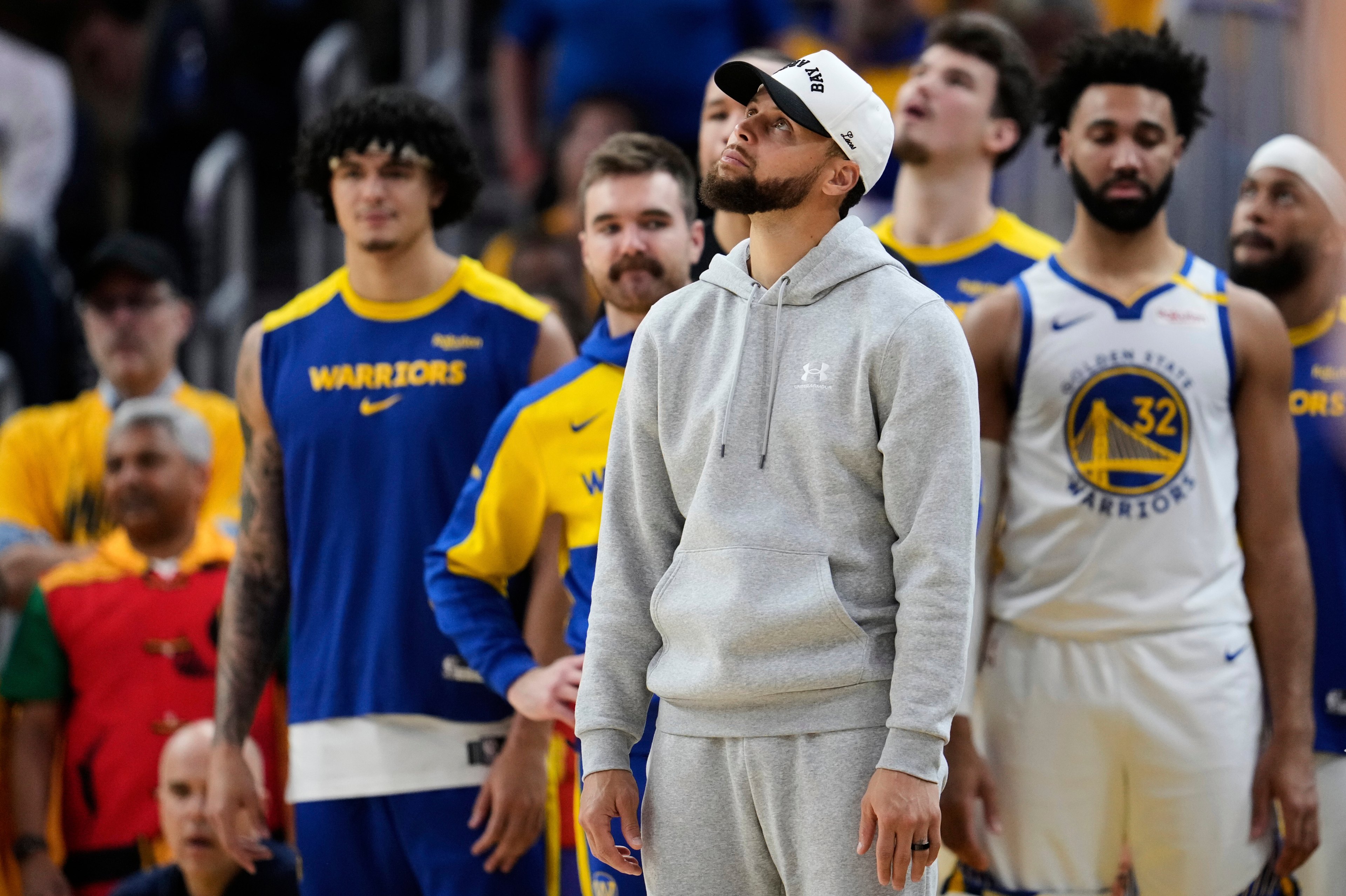 A person in a gray hoodie and white cap stands in front of basketball players wearing Golden State Warriors uniforms during a game.