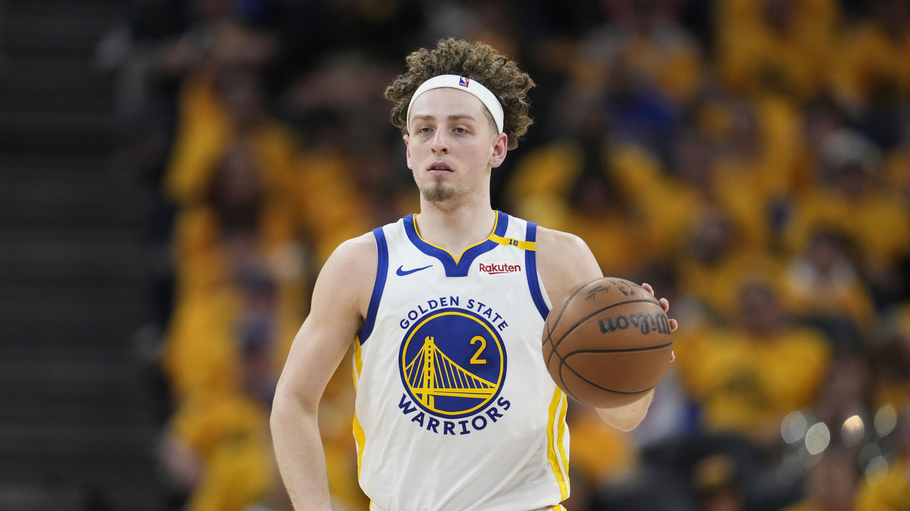 A basketball player in a Golden State Warriors jersey is dribbling a ball. He's wearing a white headband, and there's a crowd in yellow shirts in the background.