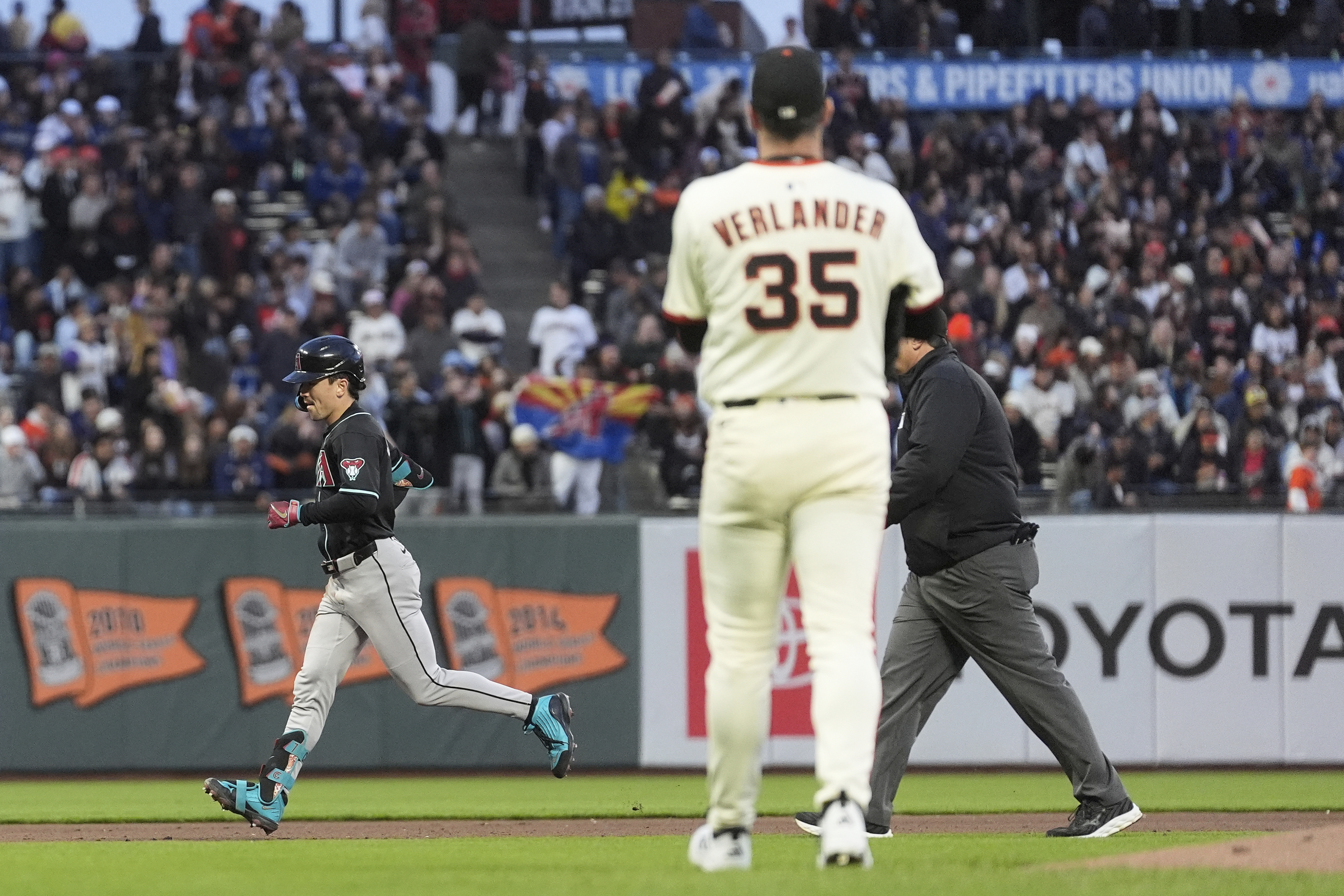 A baseball player in a black jersey runs past a pitcher in a white jersey with &quot;35&quot; on the back. An umpire and a crowd with banners are in the background.