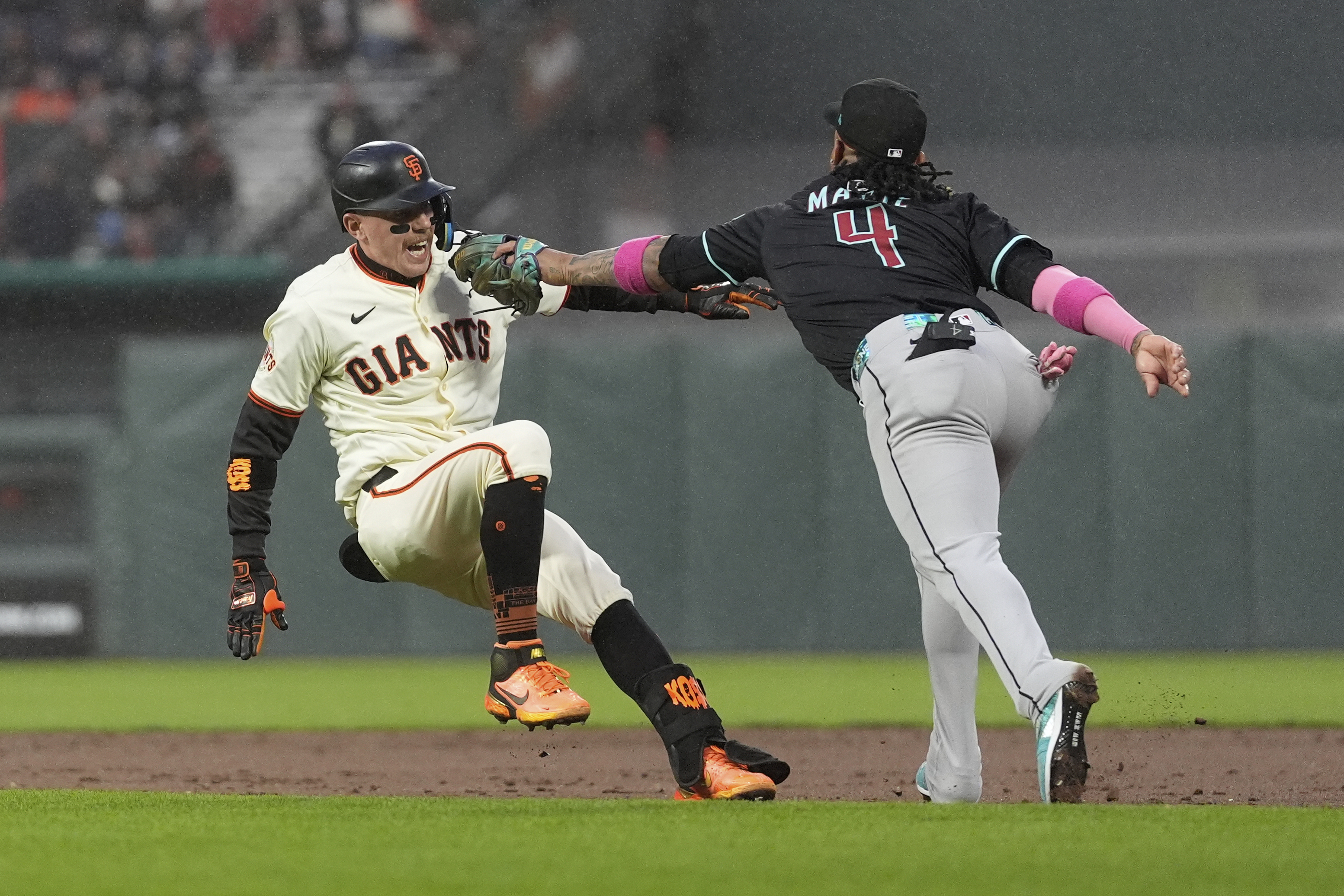 A baseball player in a white "Giants" uniform slides as an opposing player in a black and gray uniform tags him. The moment is action-packed and dynamic.