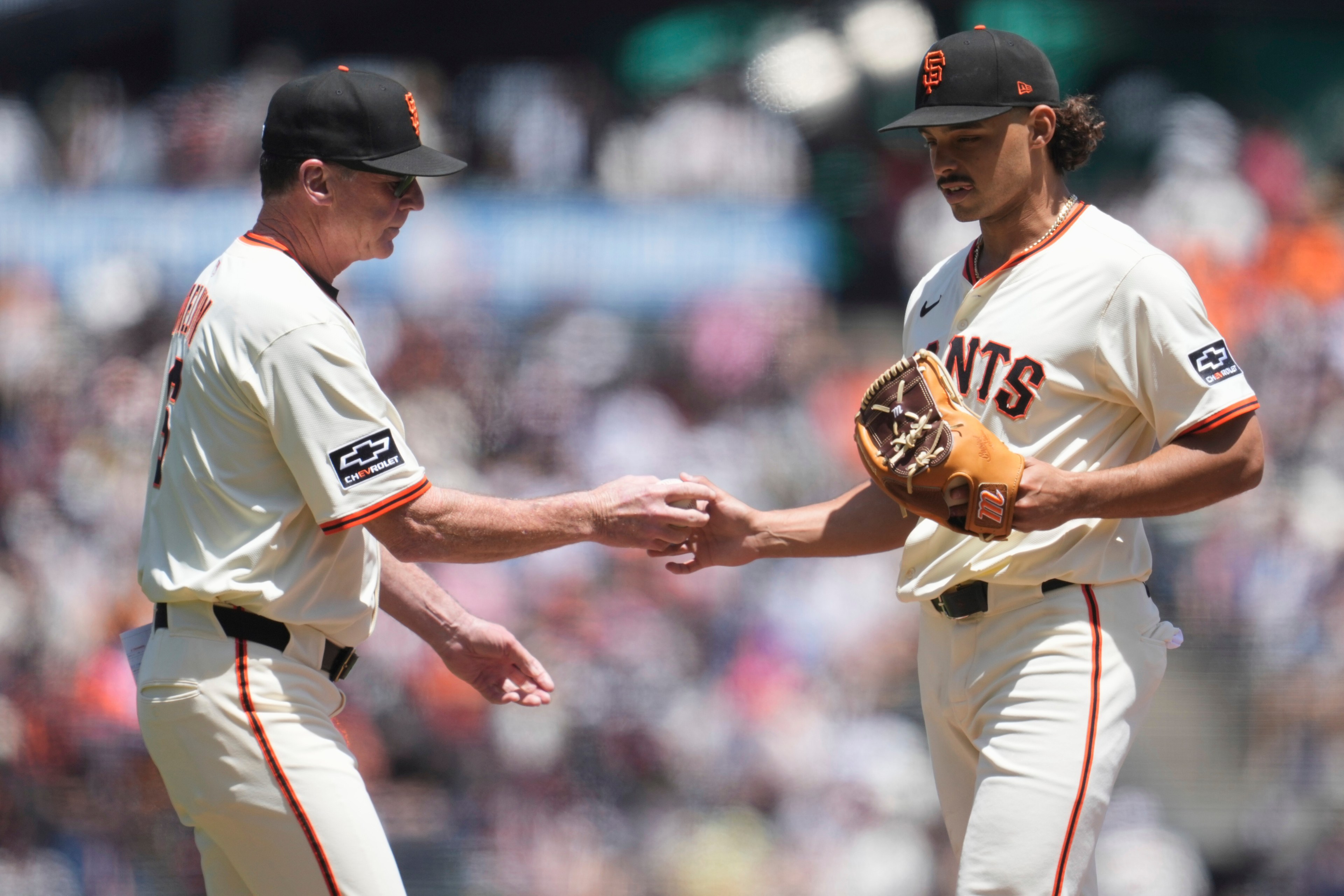 Two baseball players in white Giants uniforms stand on a field. One is handing a ball to the other, who has a glove in his hand. The background is blurred.