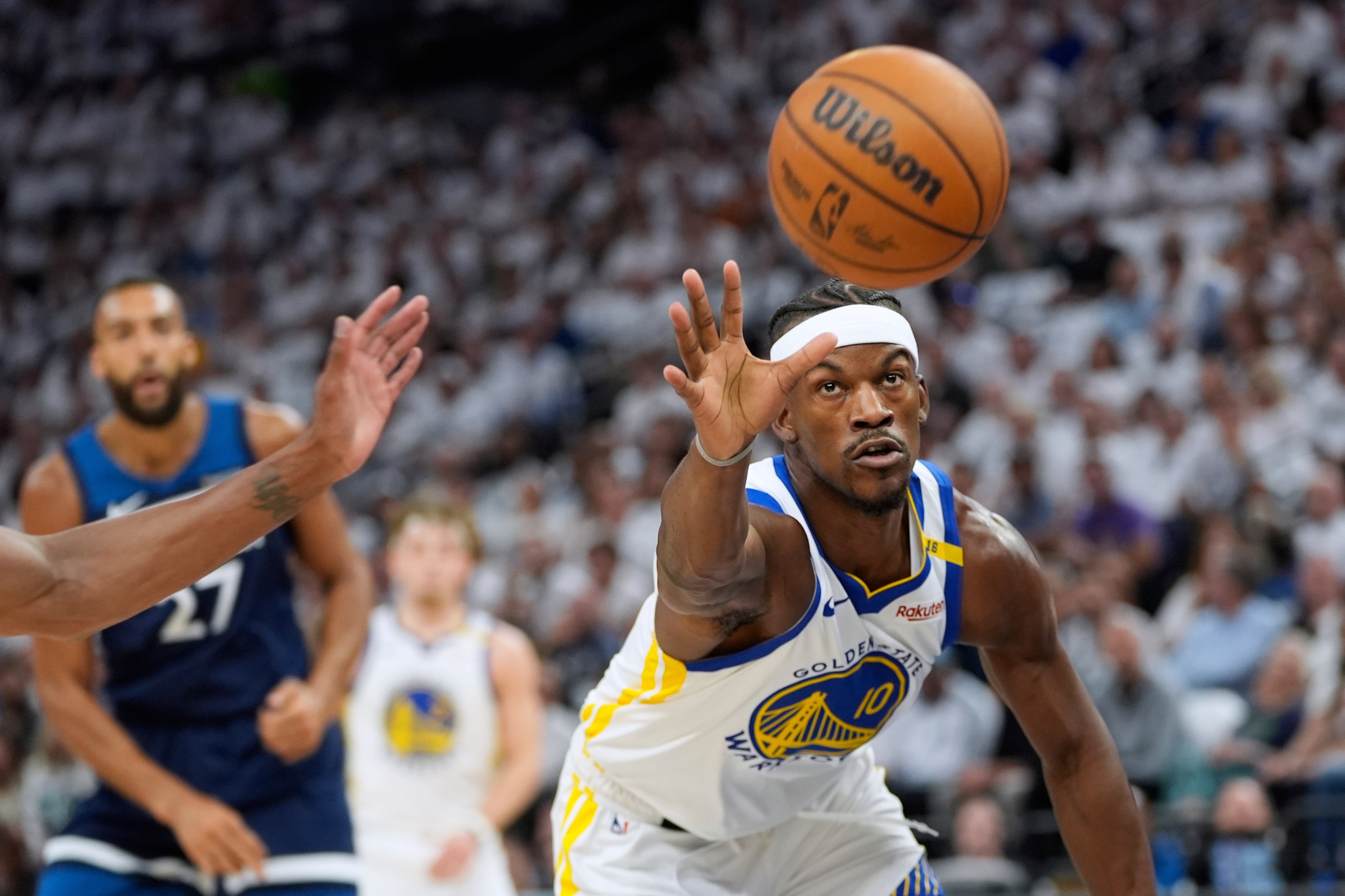 A basketball player in a white jersey, reaching for the basketball mid-air, with intense focus. Another player in blue is partly visible behind him. Crowd in the background.