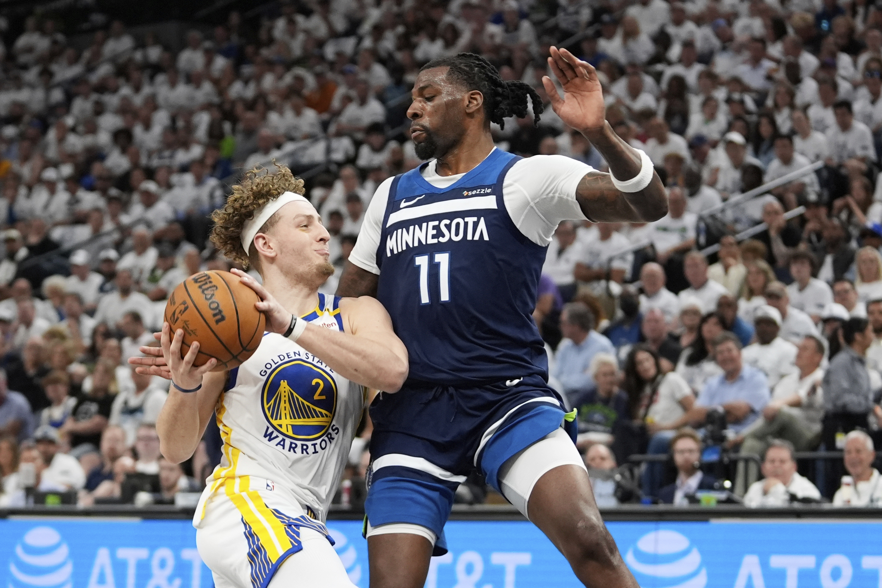 A basketball player from the Warriors is holding the ball while being closely guarded by a player from the Timberwolves. The crowd is visible in the background.
