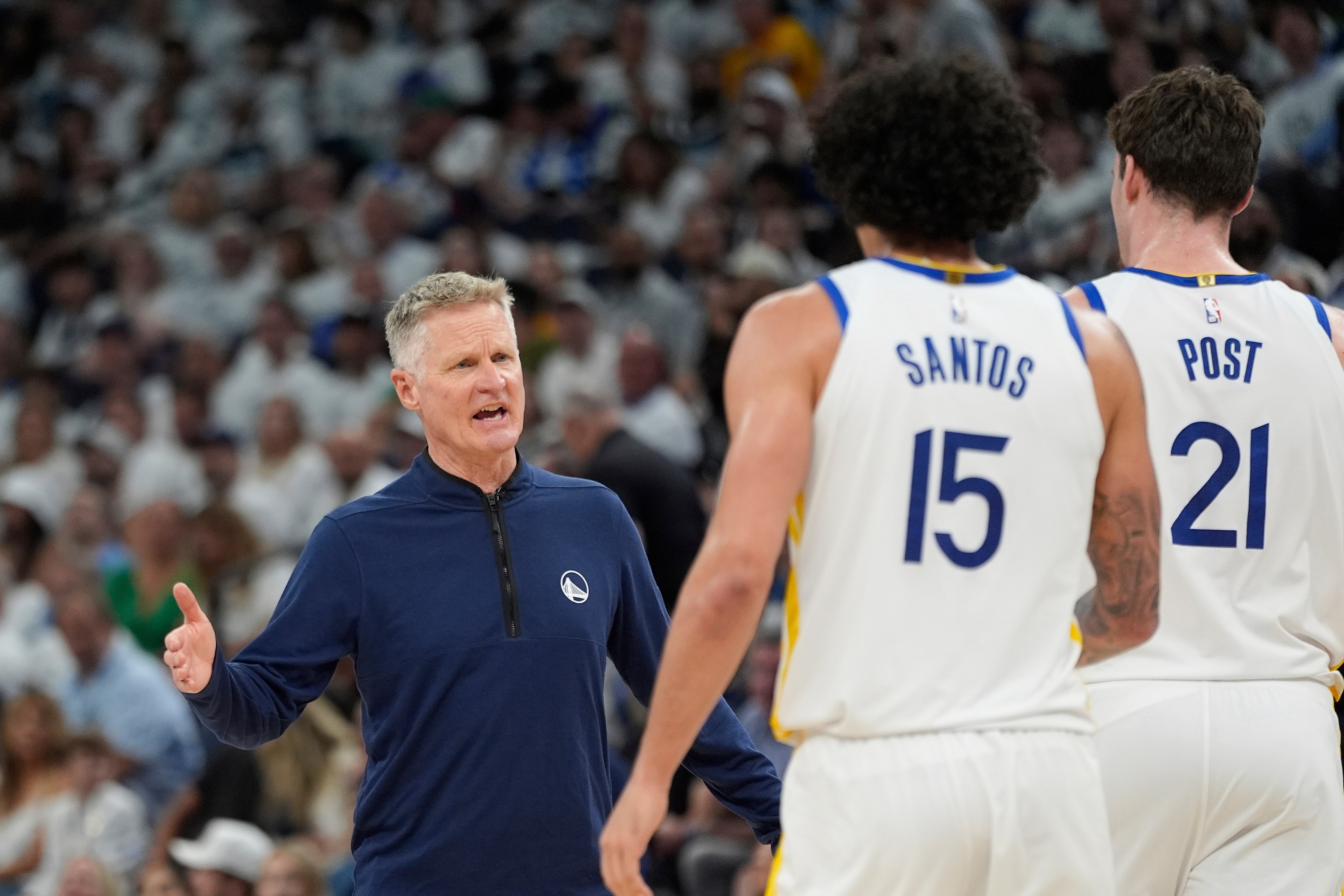 A coach gestures while talking to two basketball players wearing white jerseys numbered 15 and 21. The background shows a crowd in a sports arena.