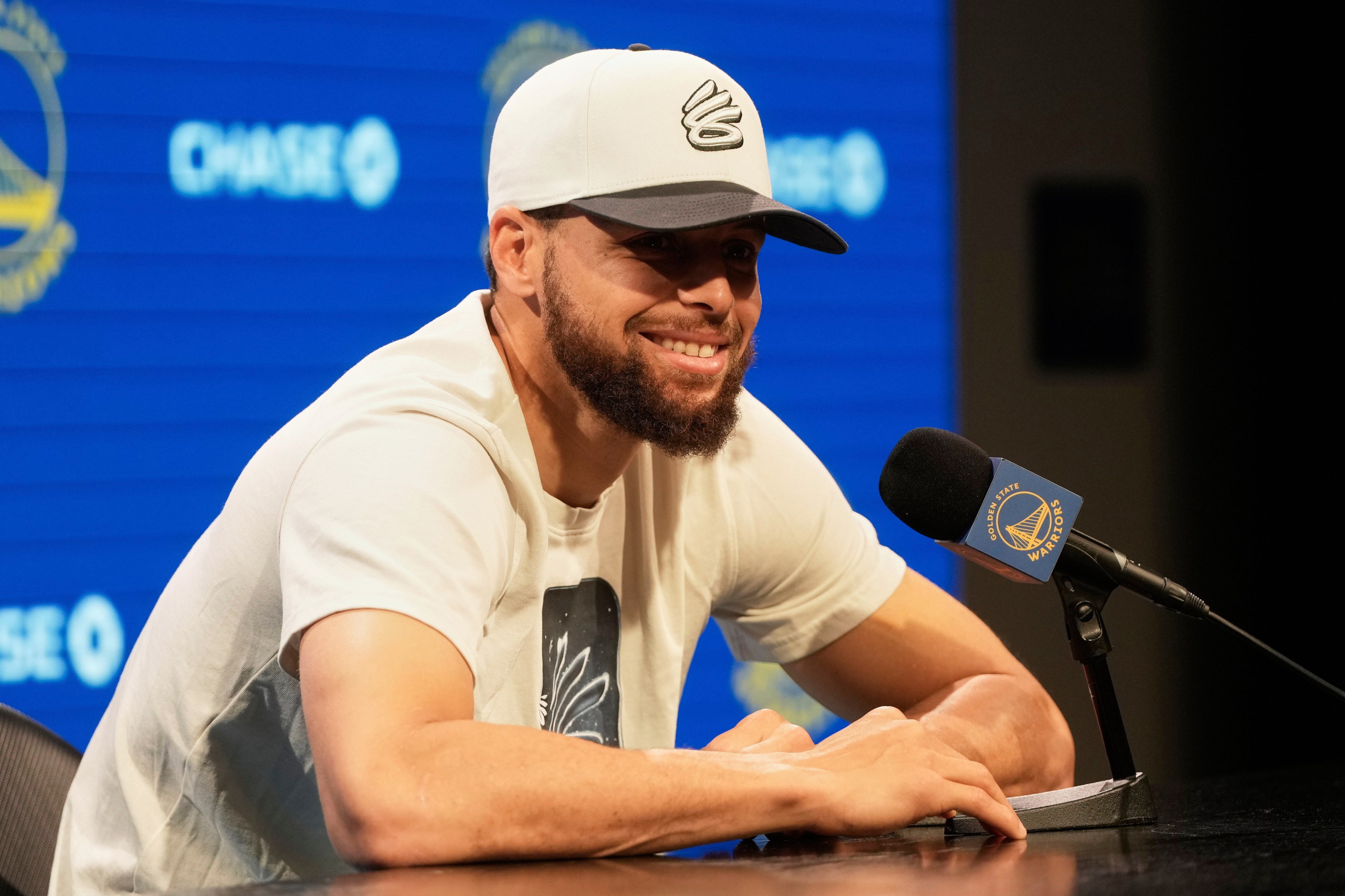 A person wearing a cap and a white t-shirt is smiling at a press conference. They're seated at a table with a microphone. The backdrop has a logo and branding.